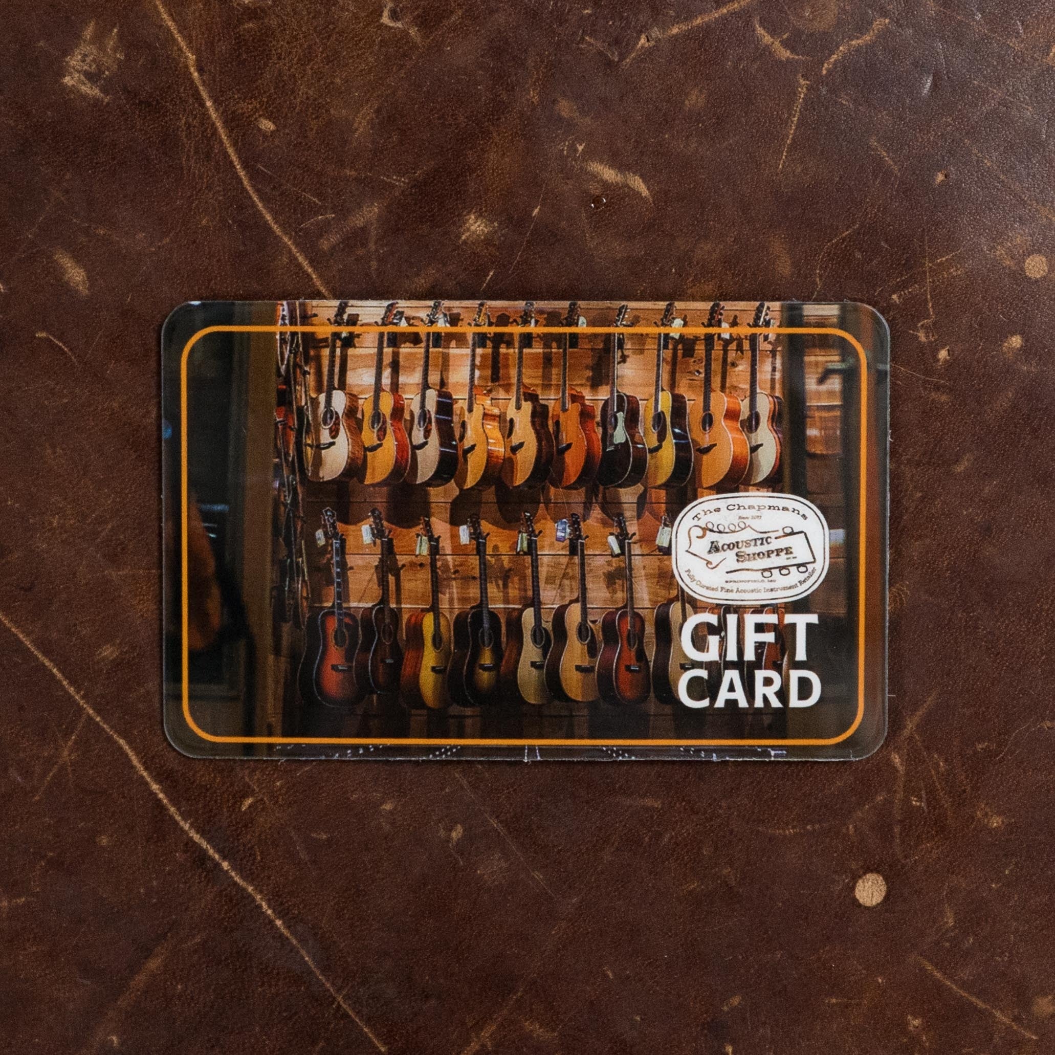 A gift card featuring the Atkin White Rice Baked Sitka/Rosewood Dreadnought Acoustic Guitar #3497 by Atkin Guitars, displayed on wood and brown leather, with a logo and the words GIFT CARD in bold white letters.