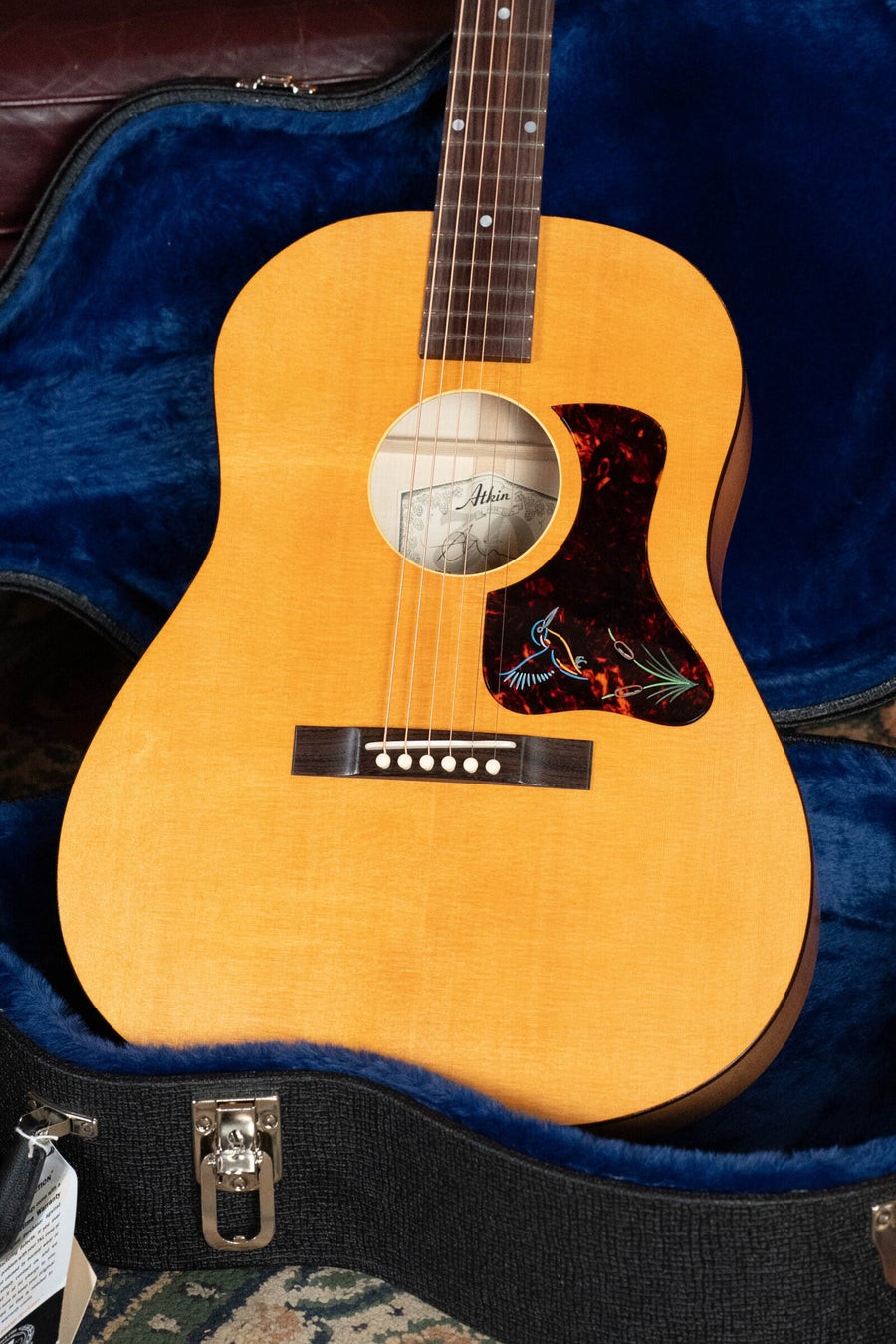 An Atkin Guitars 19 Kingfisher Sitka/Maple Dreadnought Acoustic Guitar #3892 with a natural finish sits in an open, blue velvet-lined hard case, featuring a bird pickguard and six white bridge pins.