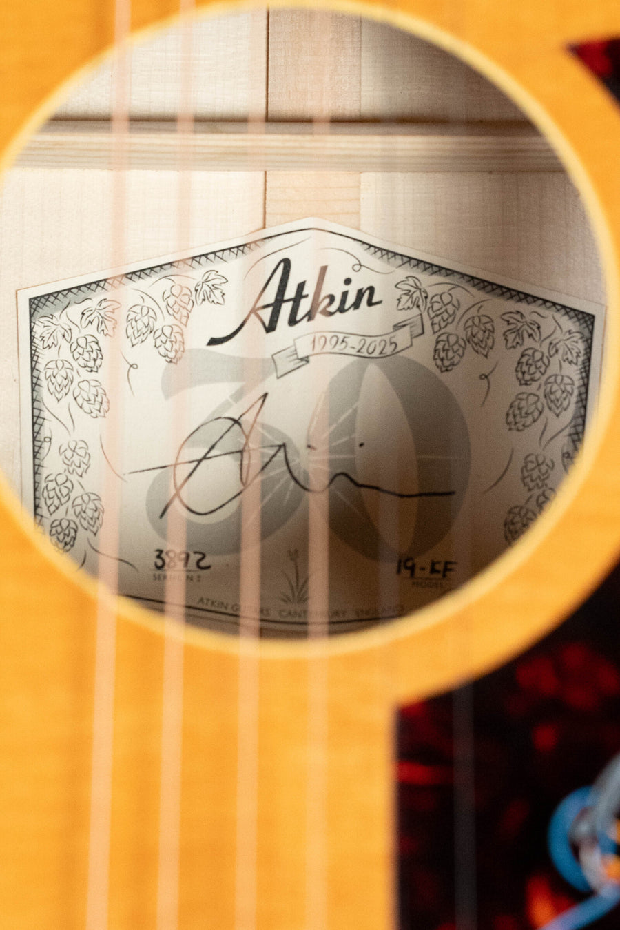 Close-up view through the soundhole of an Atkin Guitars 19 Kingfisher Sitka/Maple Dreadnought Acoustic Guitar #3892, showing a partially obscured label with handwritten brand, date, and serial number details behind the guitar strings.