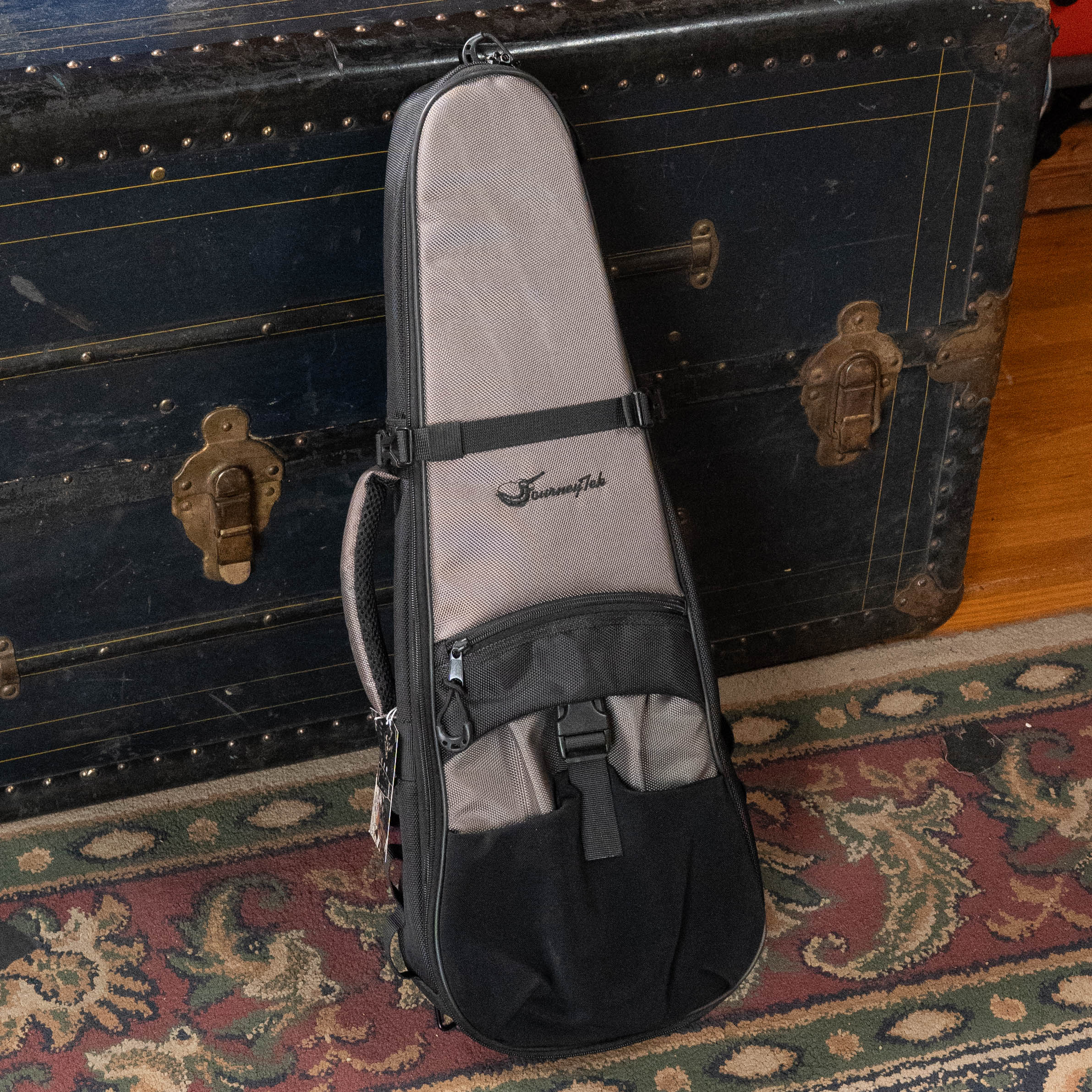 The Journey Instruments JourneyTek Concert Ukulele Case BG002 in tan and black, featuring the logo, rests against a vintage black trunk with brass hardware. A wooden floor and patterned rug highlight this protective travel case for your ukulele.