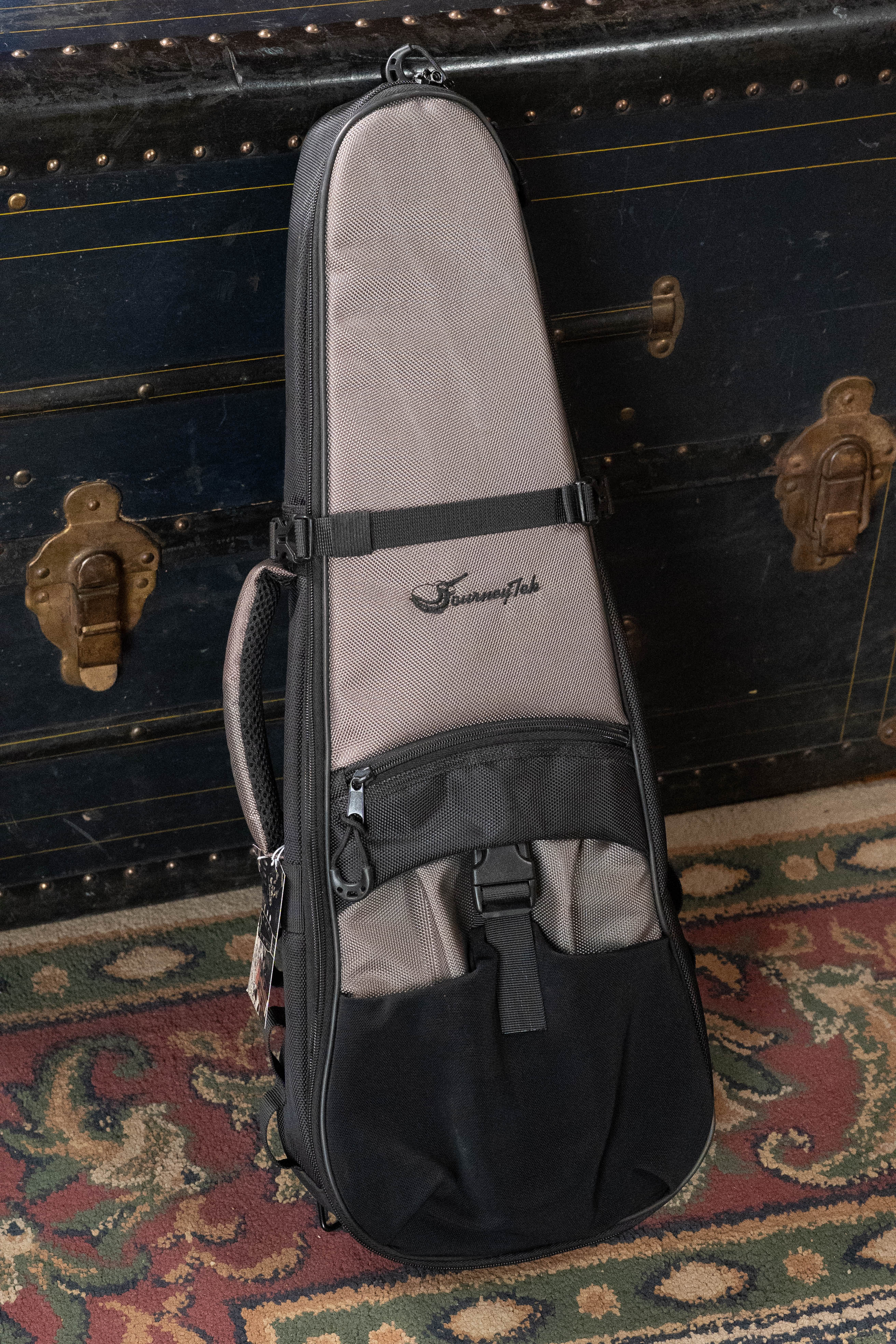 A gray and black JourneyTek Concert Ukulele Case BG002 by Journey Instruments stands upright on a patterned rug in front of a vintage black trunk with brass hardware, offering premium protection for your instrument.