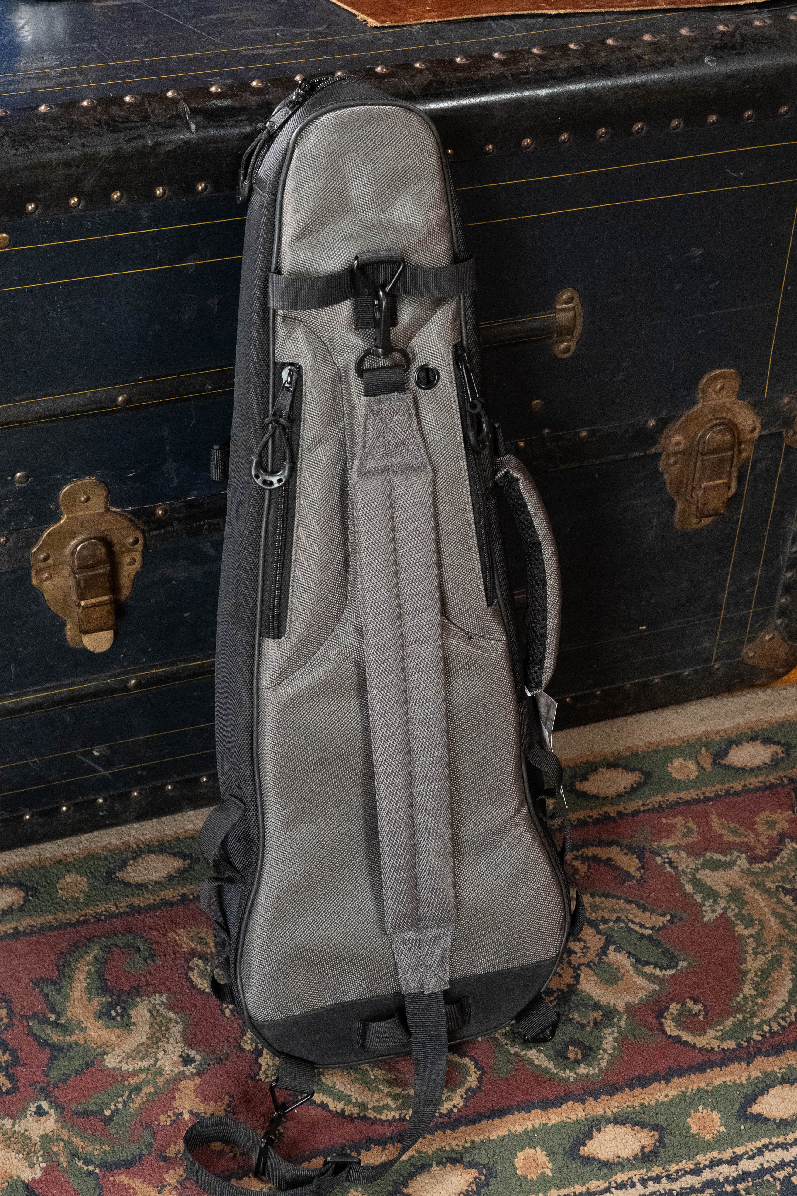 The Journey Instruments JourneyTek Concert Ukulele Case BG002, a gray padded backpack with shoulder straps, leans against a vintage trunk on a red, green, and beige patterned rug for stylish instrument protection.