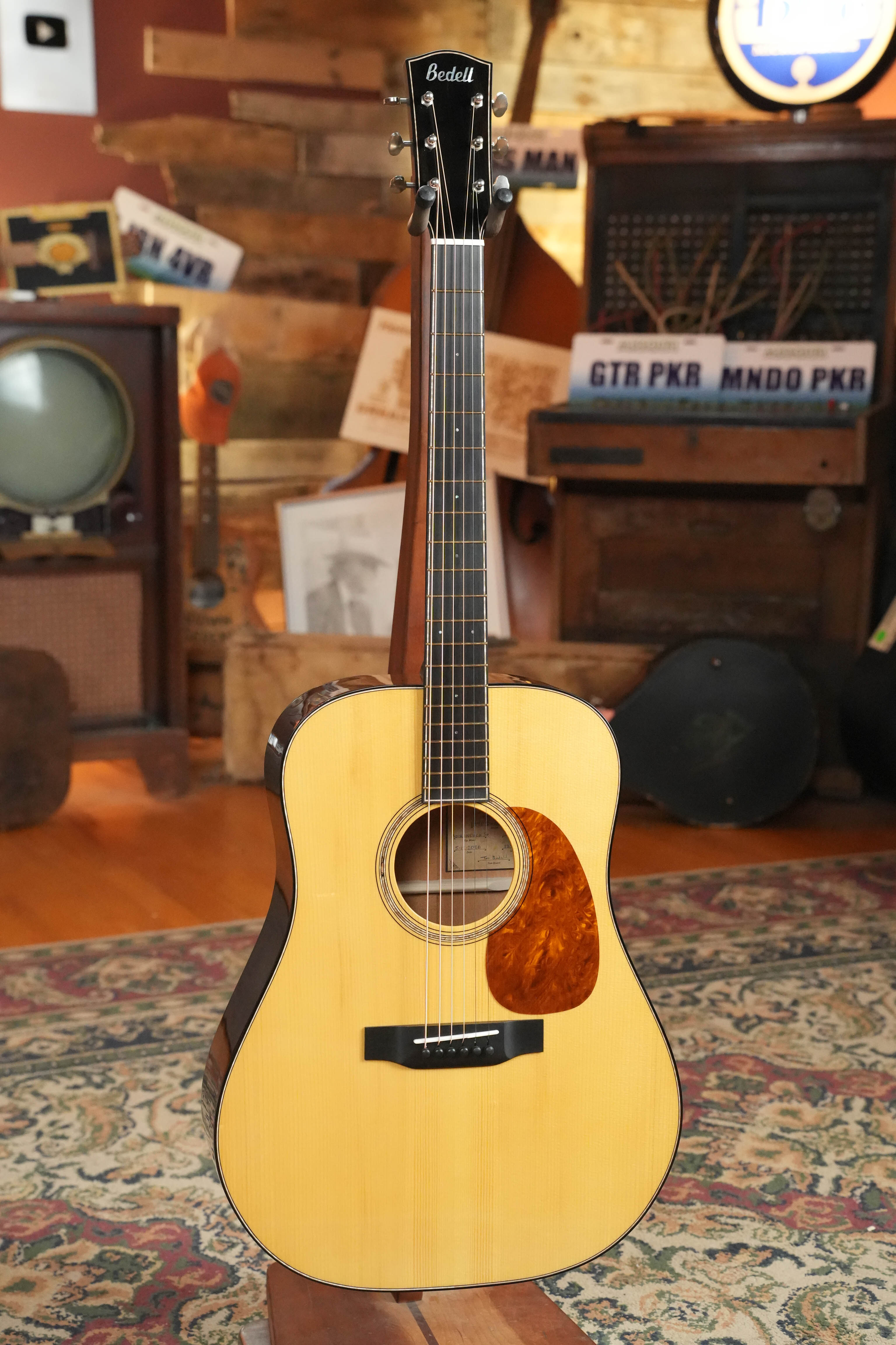 The Bedell Guitars Special Edition 1940 Dreadnought Narrow Nut Acoustic Guitar #5004, featuring a light wood body, Adirondack spruce top, dark sides, and tortoiseshell pickguard, stands upright on a patterned rug amid vintage decor.