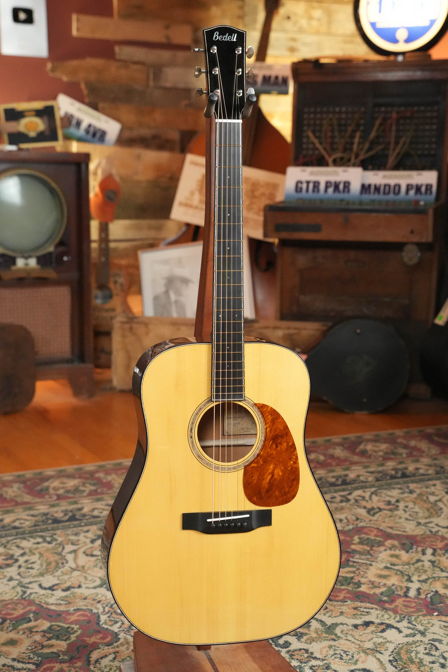 The Bedell Guitars Special Edition 1940 Dreadnought Narrow Nut Acoustic Guitar #5004, featuring a light wood body, Adirondack spruce top, dark sides, and tortoiseshell pickguard, stands upright on a patterned rug amid vintage decor.
