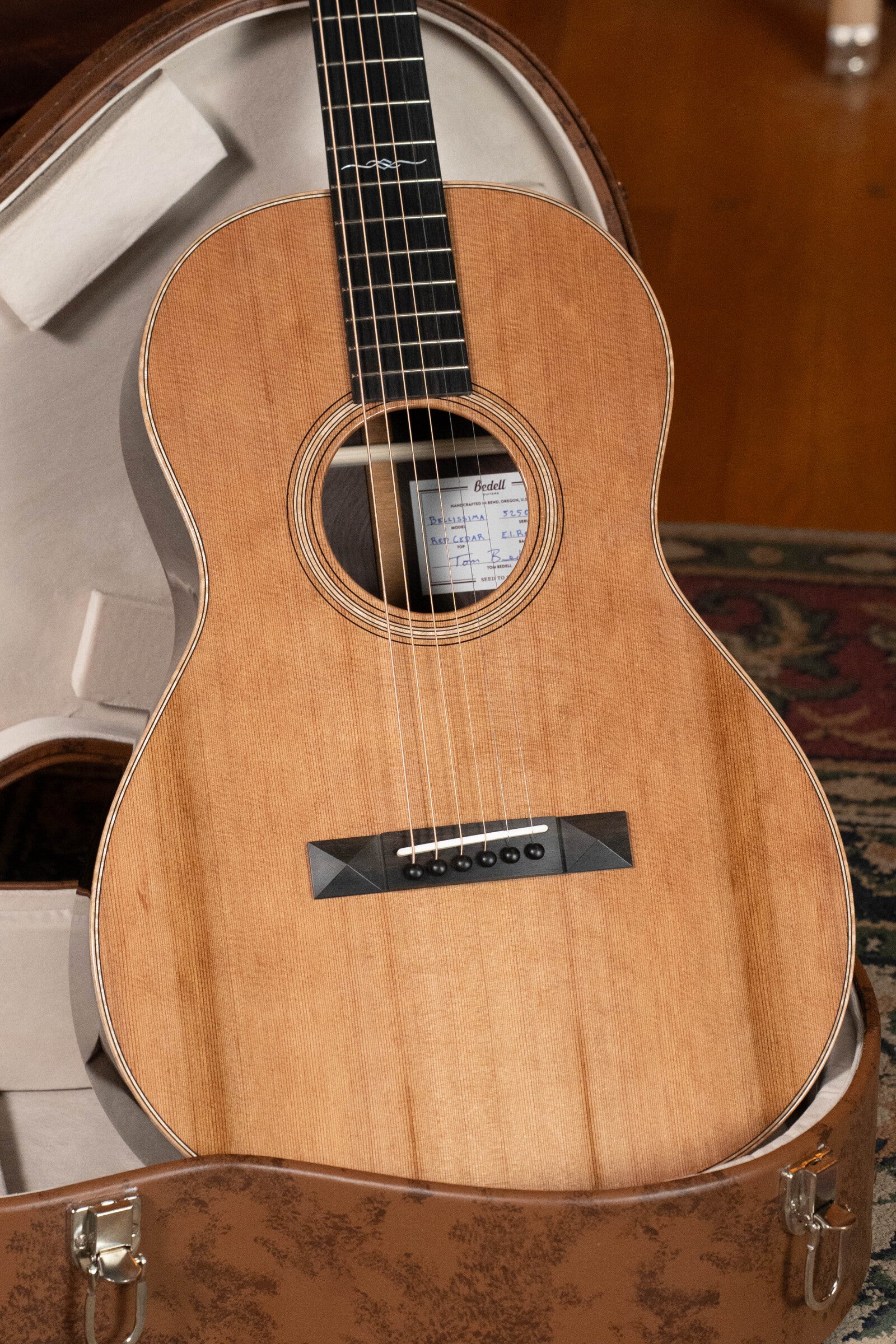A Bedell Guitars Angelica Bellissima Red Cedar/Rosewood Parlor Guitar w/K&K Pure Mini Pickup (#5002) with a natural finish rests in a tan, plush-lined hard case on a wooden floor near a patterned rug.