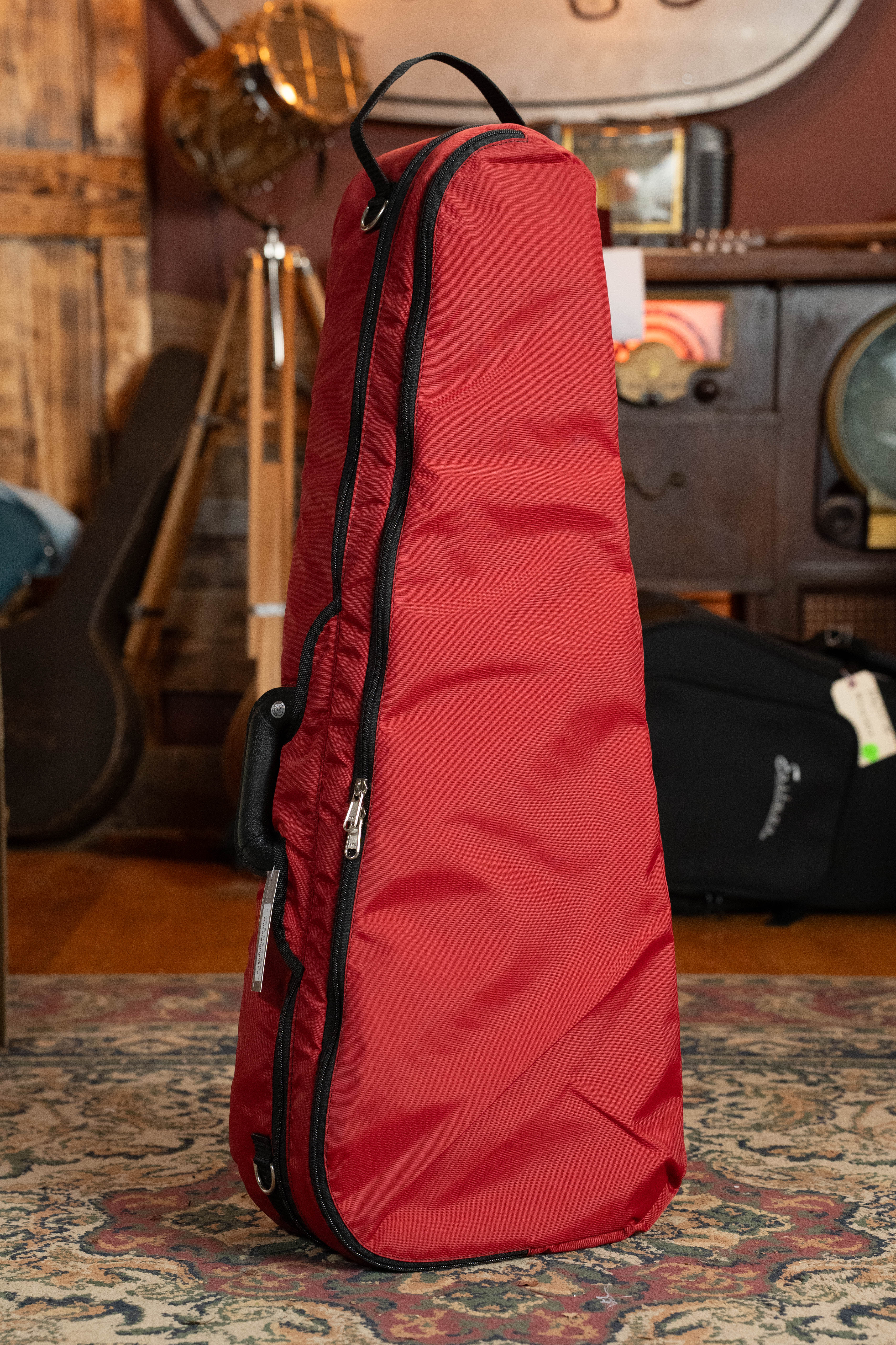 The Bobelock Arrow Red/Gray F-Style Fiberglass Mandolin Case by Howard Core stands upright on a patterned rug in a cozy room with wooden decor and vintage instruments in the background.