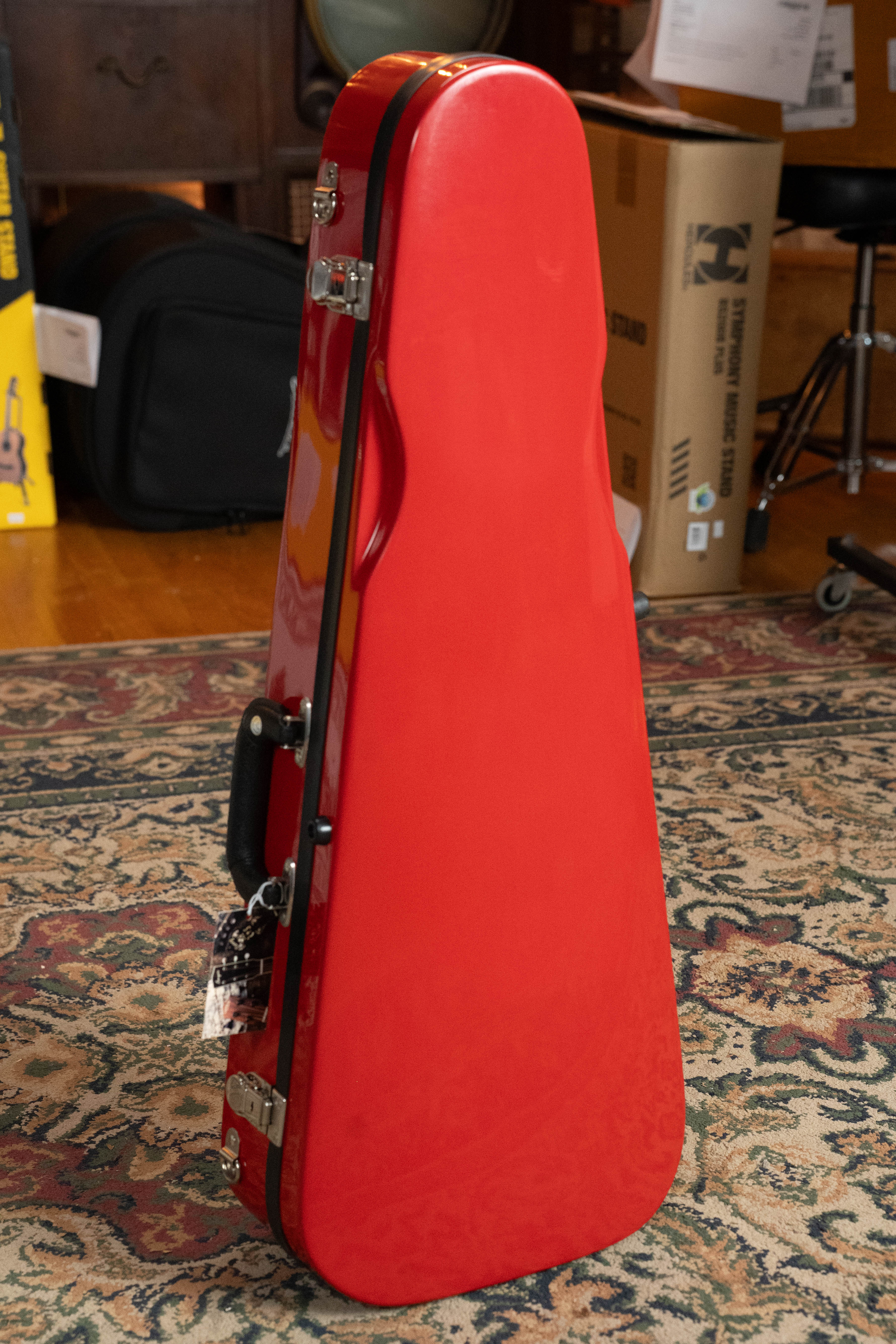 The Howard Core Bobelock Arrow Red/Gray F-Style Fiberglass Mandolin Case, featuring silver latches and a black handle, stands upright on a patterned rug among boxes and furniture in the room.