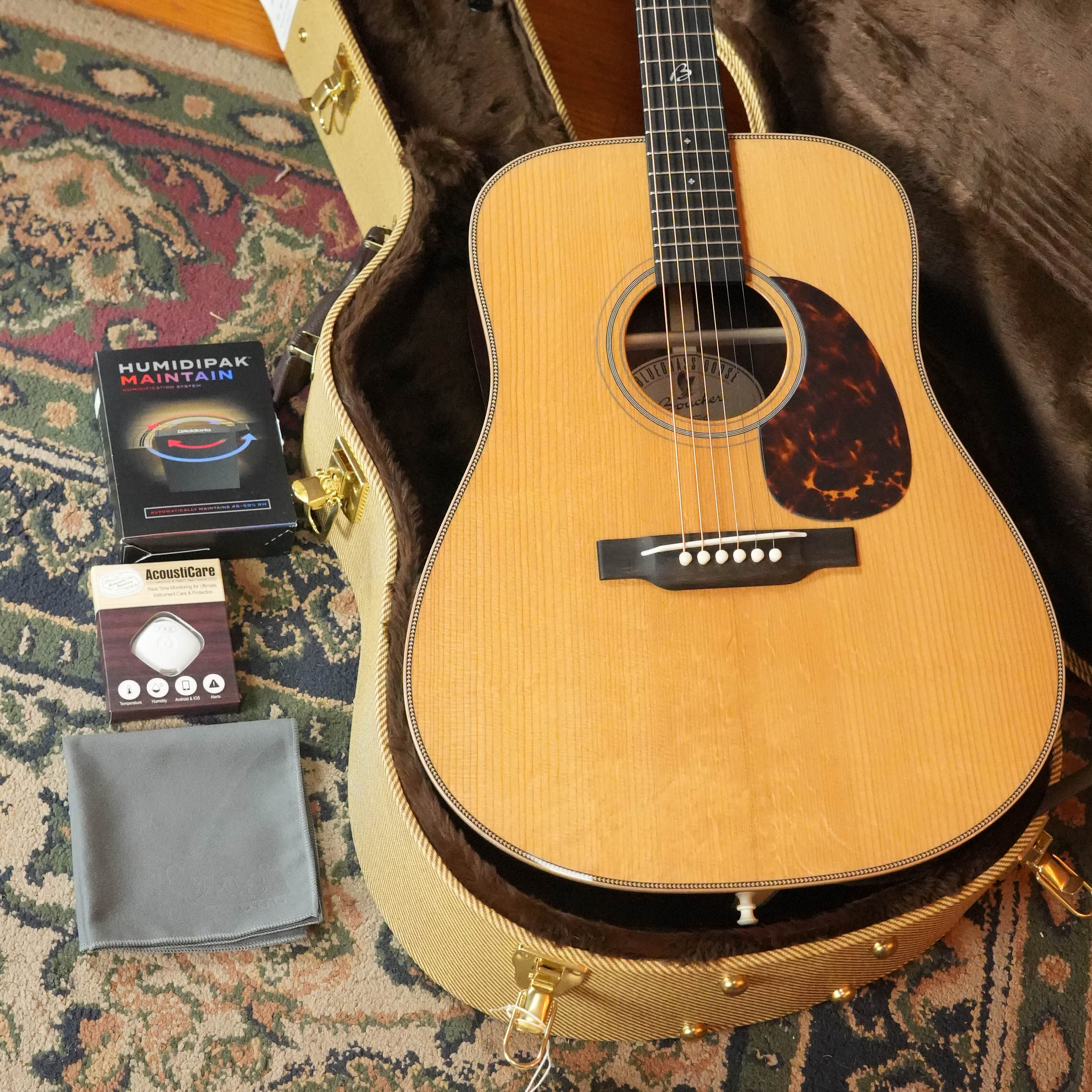 A Boucher Guitars BG-152-G Adirondack/Rosewood Bluegrass Goose Dreadnought Acoustic Guitar (#1098) sits in an open, plush-lined hard case on a patterned rug, with a Humidipak system, string winder, and cleaning cloth nearby.
