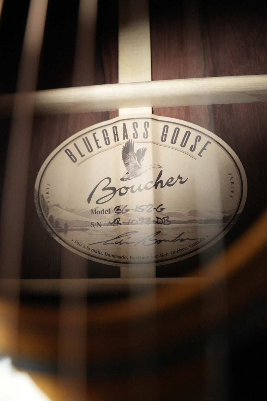 Close-up of a Boucher Guitars BG-152-G Adirondack/Rosewood Bluegrass Goose Dreadnought Acoustic Guitar #1098 label, featuring a goose illustration and serial number, photographed through the strings.