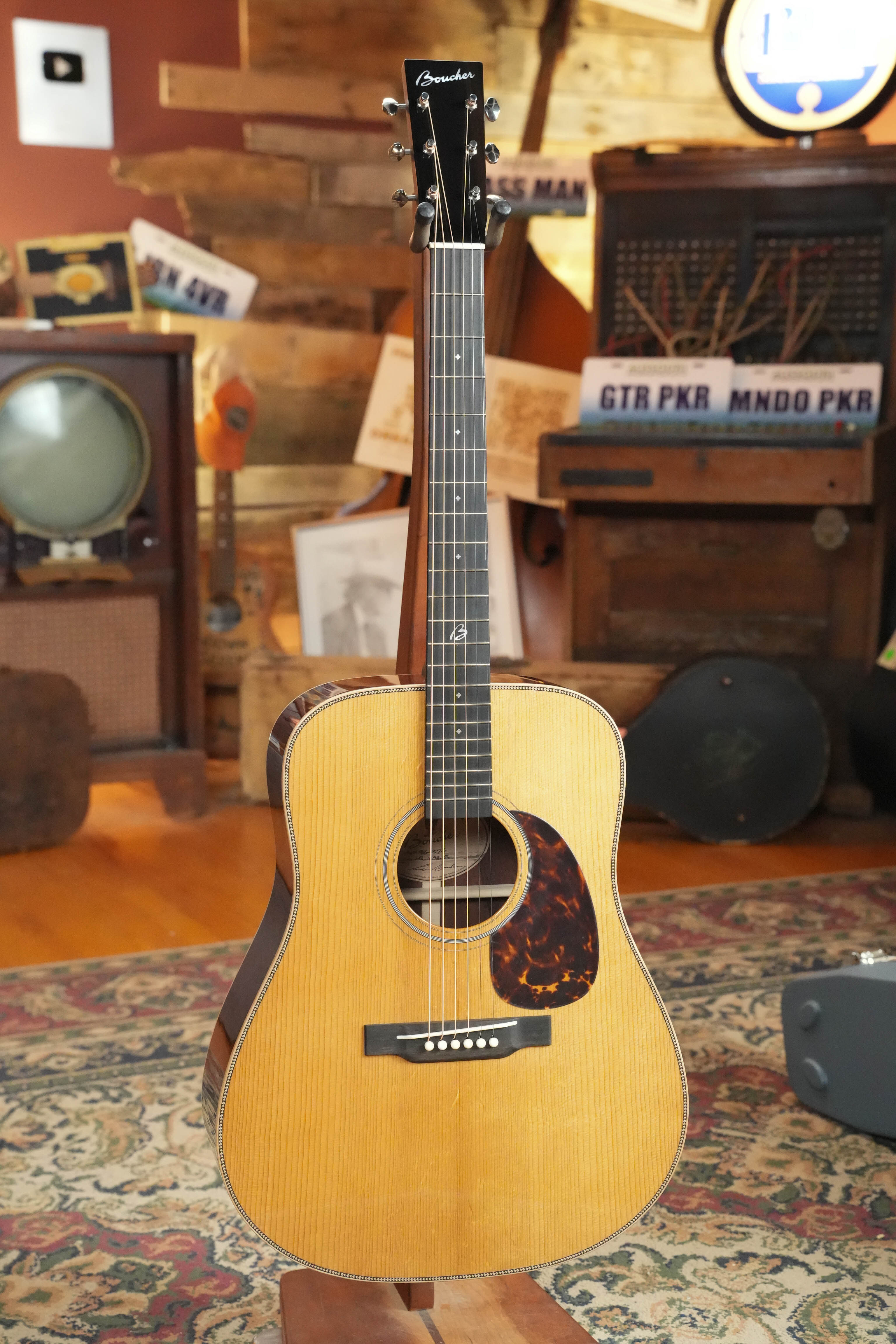 A Boucher Guitars BG-152-G Adirondack/Rosewood Bluegrass Goose Dreadnought Acoustic Guitar #1098 with a natural finish stands upright on a patterned rug in a cozy, vintage-themed room with wooden walls and retro decor.