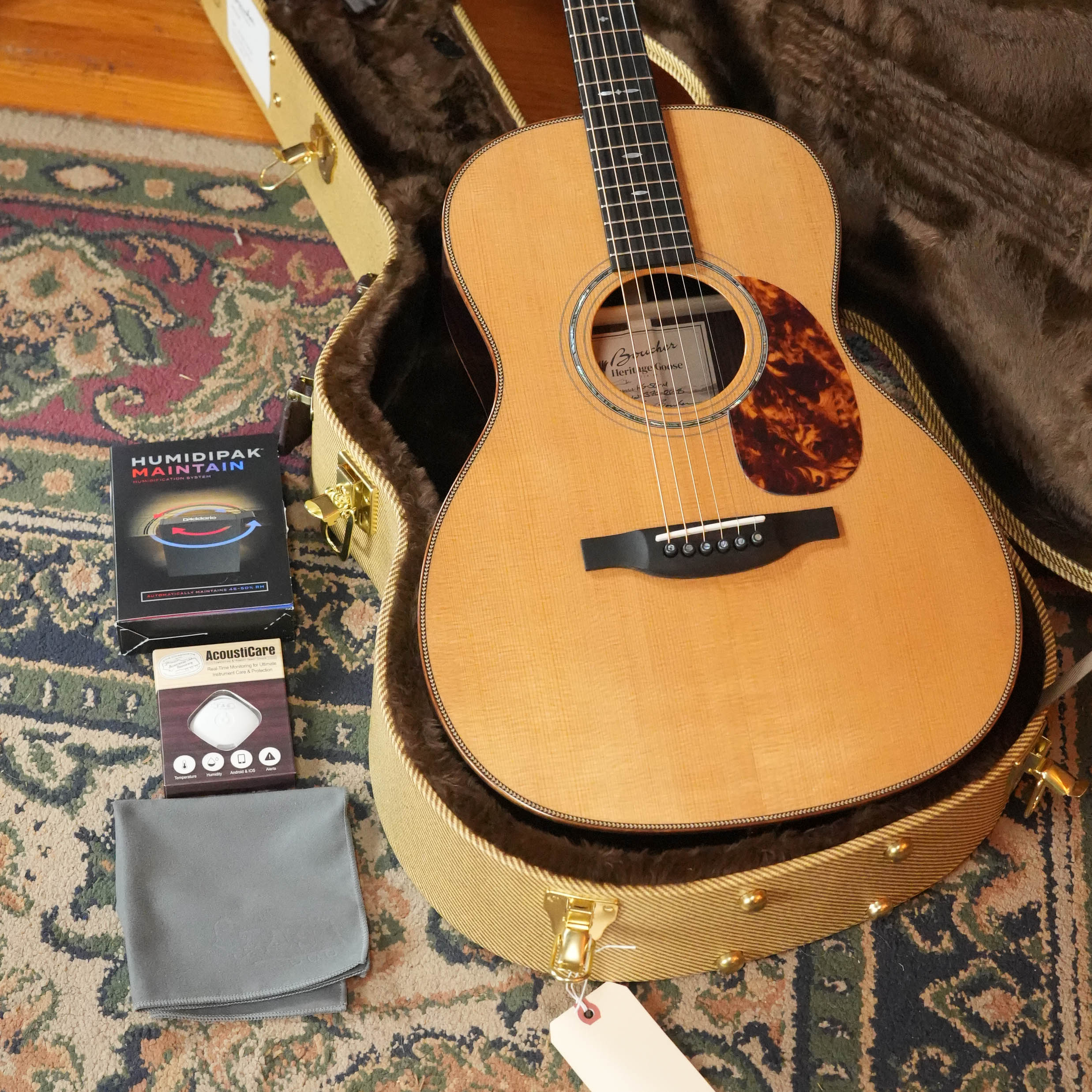The Acoustic Shoppe’s Boucher HG-56-M Adirondack/Rosewood OOO Acoustic Guitar #1370 sits in an open, plush-lined hard case with a maintenance kit, acoustic guitar strings, and a gray cleaning cloth on a colorful patterned rug.