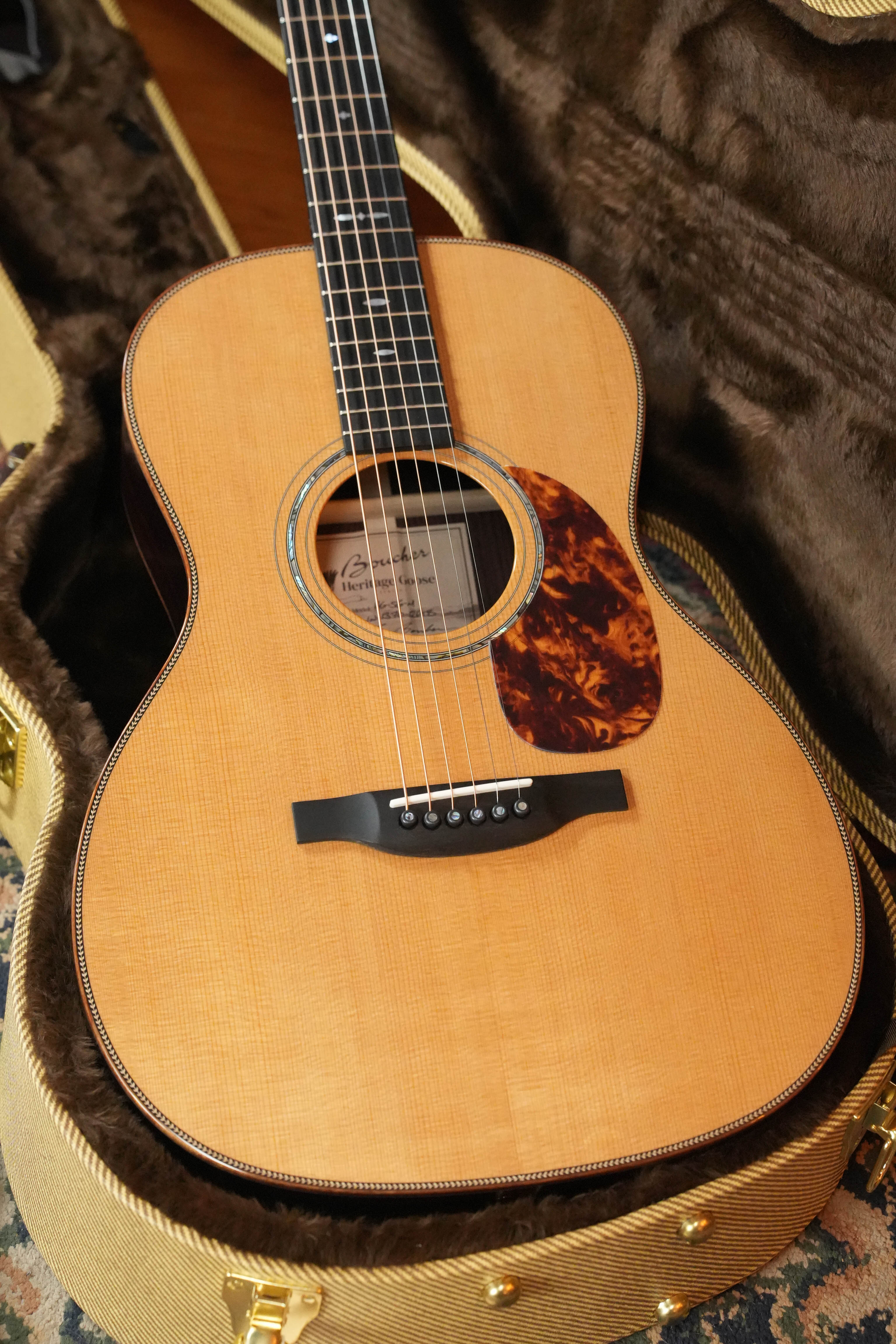 The Acoustic Shoppe’s Boucher HG-56-M Adirondack/Rosewood OOO Acoustic Guitar #1370, with a light wood top, tortoiseshell pickguard, and black bridge, rests in a plush brown-lined hard case—showcasing artisan luthiery.