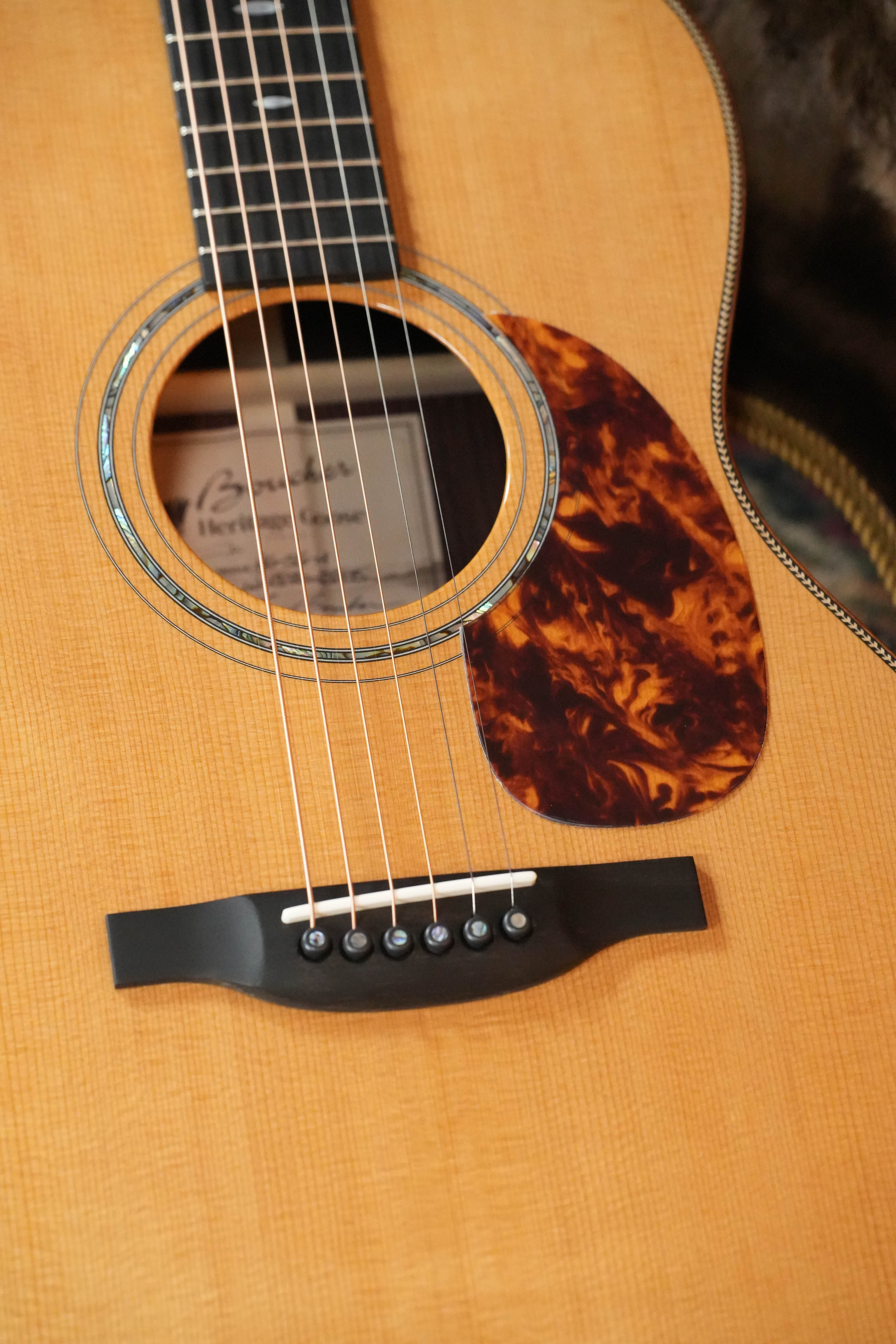 Close-up of the Boucher HG-56-M Adirondack/Rosewood OOO Acoustic Guitar #1370’s body from The Acoustic Shoppe, featuring its strings, bridge, sound hole, and tortoiseshell-style pickguard on a light wood finish.