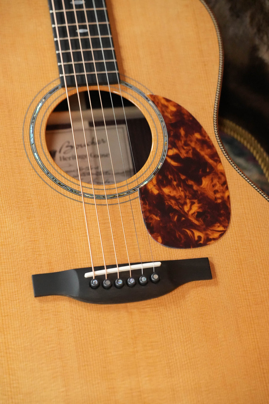 Close-up of the Boucher HG-56-M Adirondack/Rosewood OOO Acoustic Guitar #1370’s body from The Acoustic Shoppe, featuring its strings, bridge, sound hole, and tortoiseshell-style pickguard on a light wood finish.