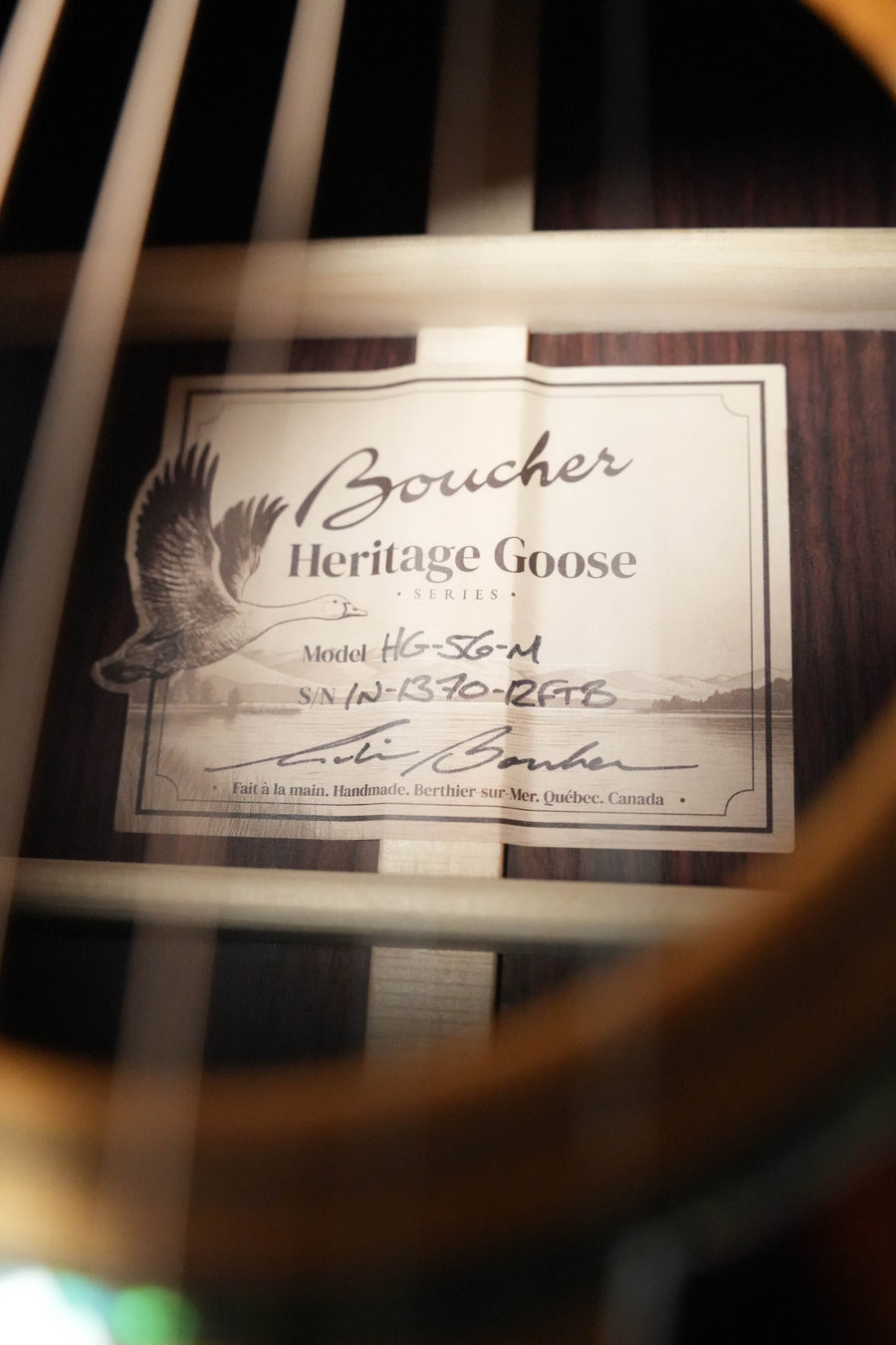 Close-up of a Boucher HG-56-M Adirondack/Rosewood OOO Acoustic Guitar #1370 label inside the soundhole at The Acoustic Shoppe, showing model, serial number, signature, and part of its torrefied Adirondack top with visible strings.