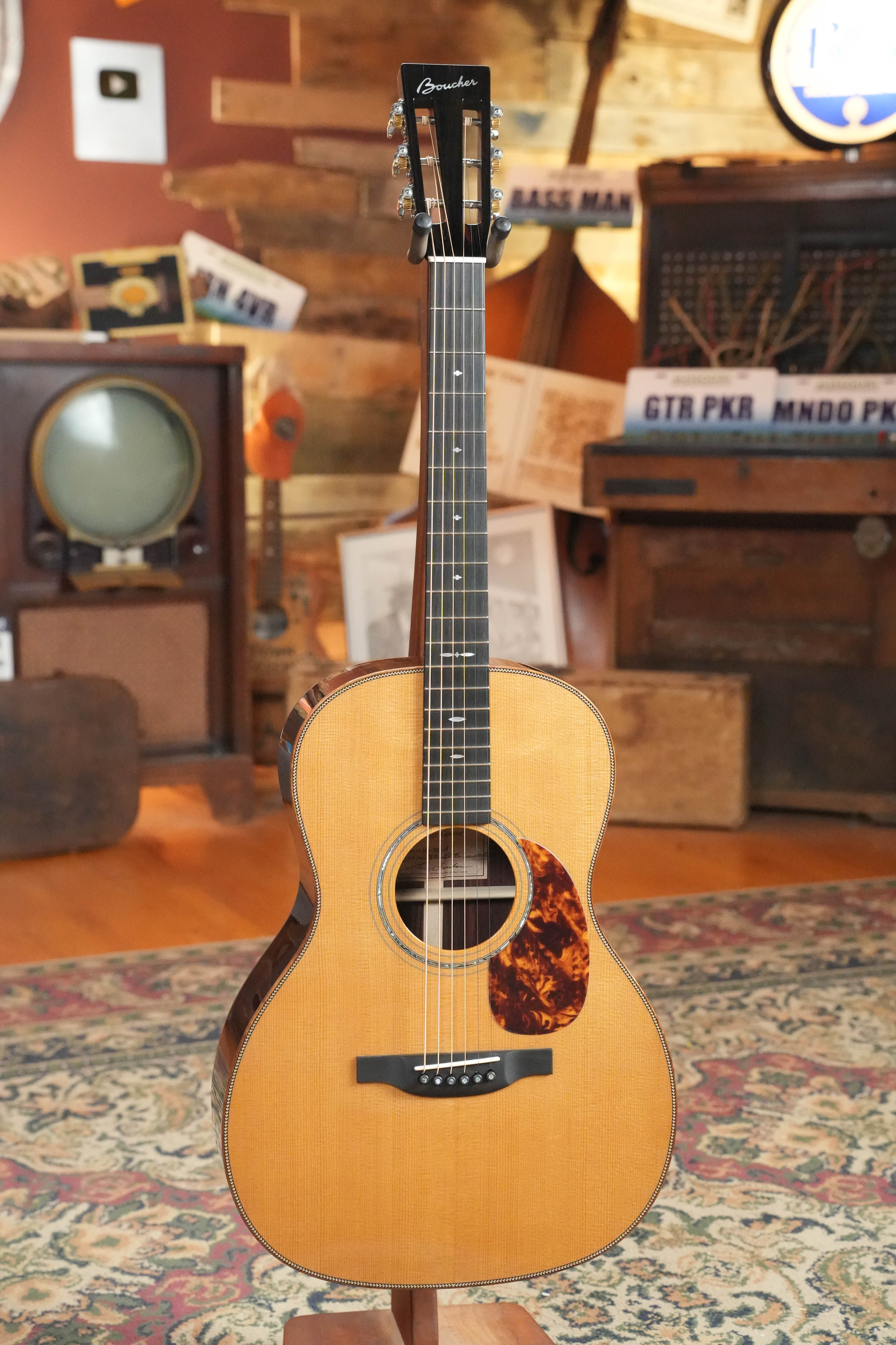 The Acoustic Shoppe’s Boucher HG-56-M Adirondack/Rosewood OOO Acoustic Guitar #1370, featuring a torrefied Adirondack spruce top and natural wood finish, stands upright on a patterned rug amid vintage decor and an old TV.