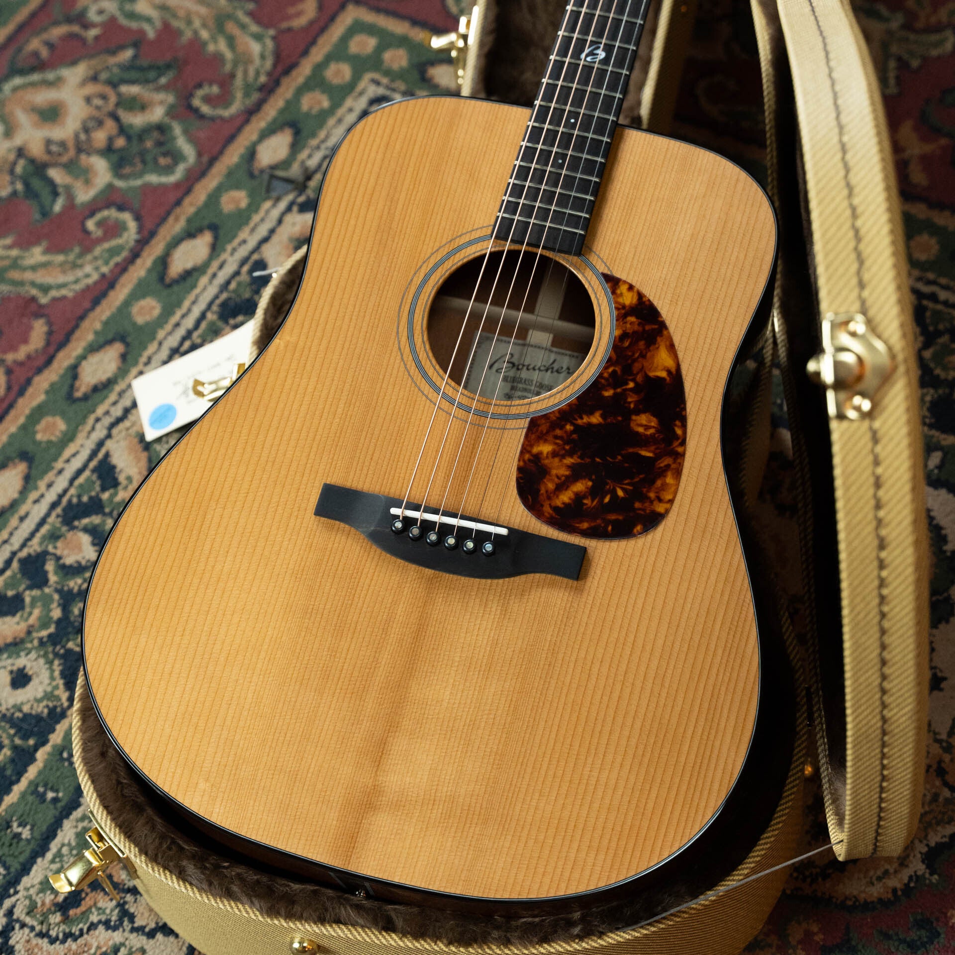 A Boucher Guitars BG-42T-G Bluegrass Goose Series torrefied master grade Adirondack/mahogany dreadnought acoustic guitar (#1035), with a natural finish and tortoiseshell pickguard, rests in an open hard case lined with dark, soft material on a patterned rug.