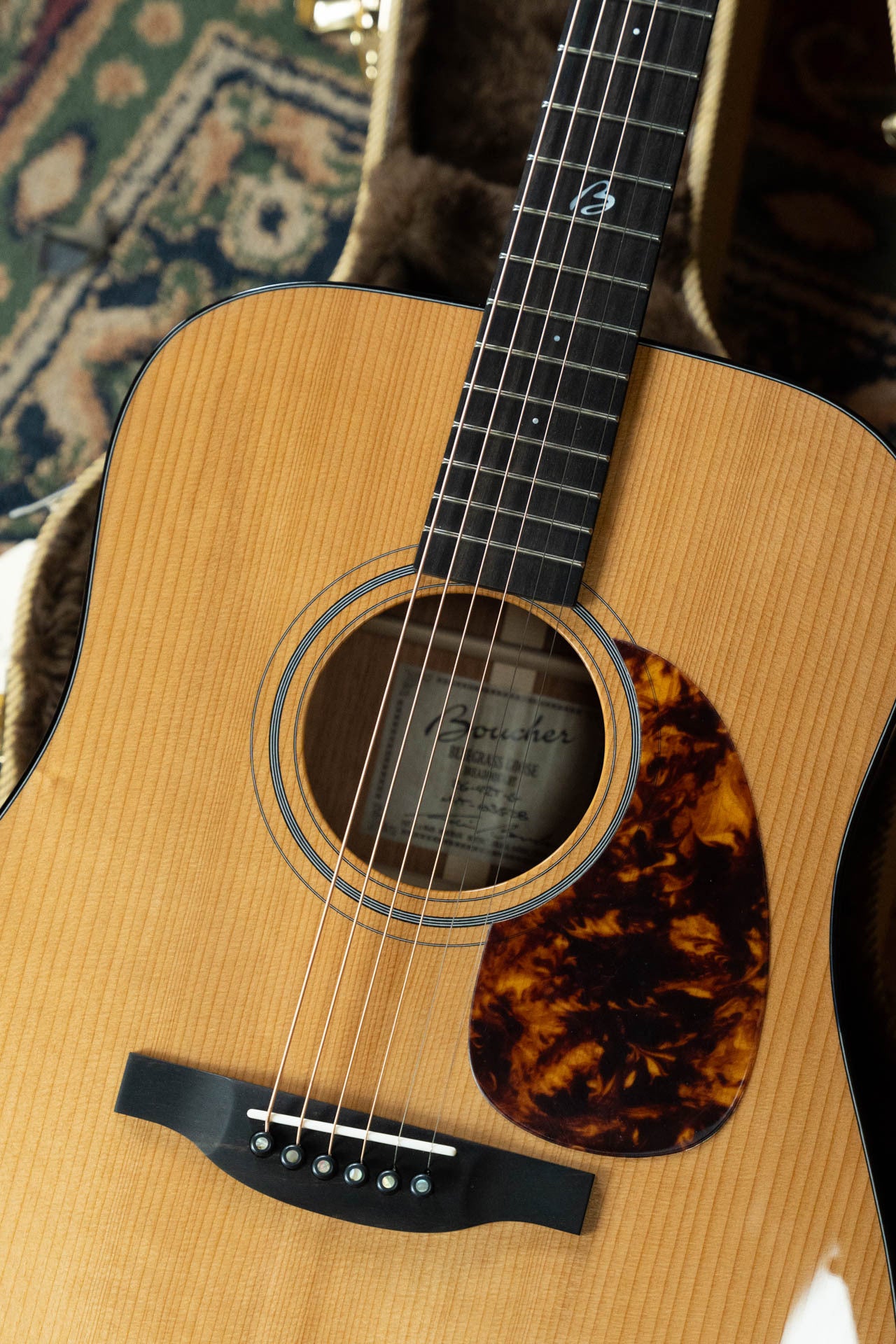 A close-up of the Boucher Guitars BG-42T-G Bluegrass Goose dreadnought acoustic guitar—ALL torrefied master grade Adirondack/mahogany—with a tortoiseshell pickguard, resting in its case on a patterned rug, ideal for bluegrass fans.