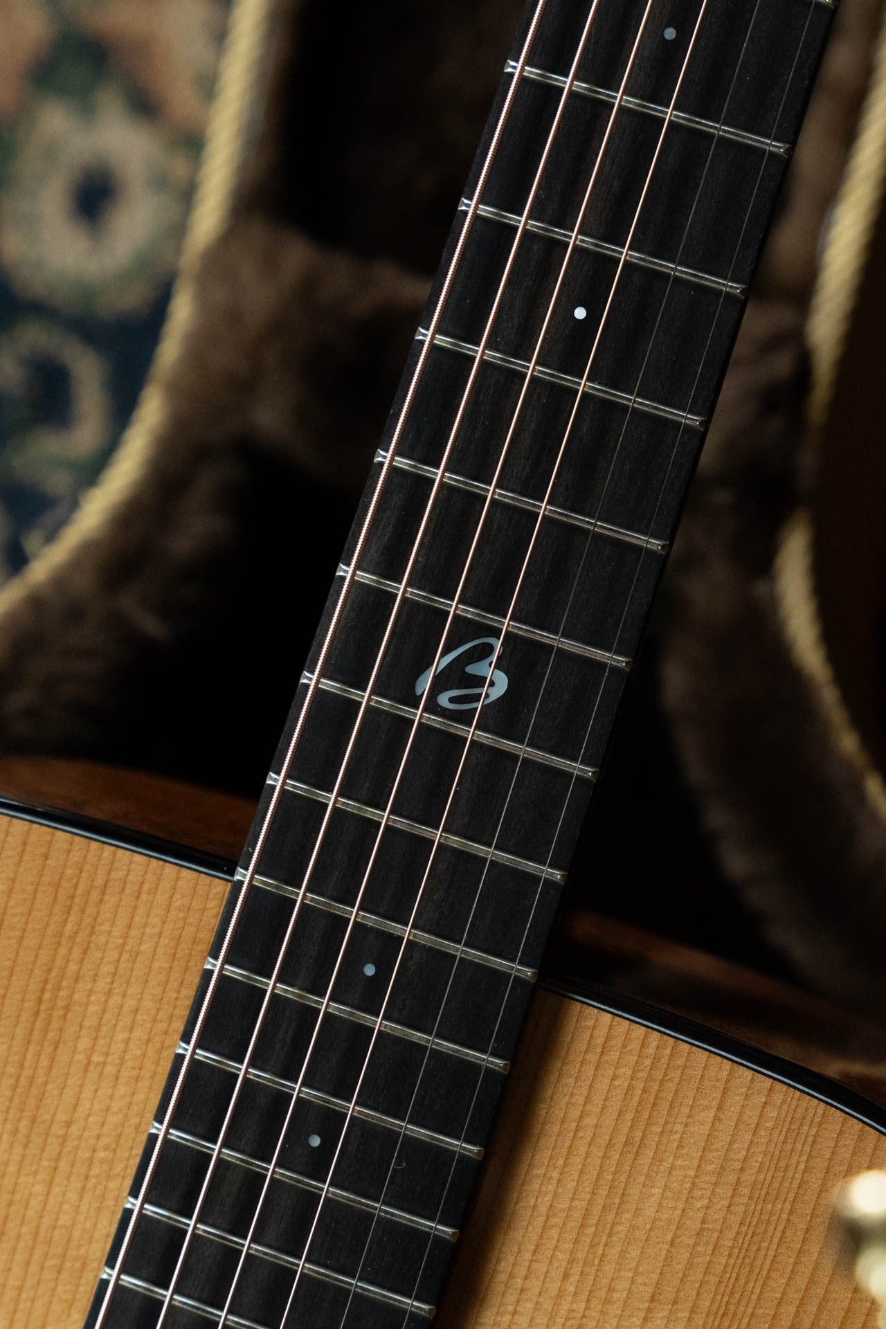 A close-up of the Boucher Guitars BG-42T-G Bluegrass Goose Series #1035 shows its torrefied Adirondack/Mahogany neck, wood grain, metal frets, dot inlays, and a unique 12th fret symbol; a soft-lined case is partly visible behind.