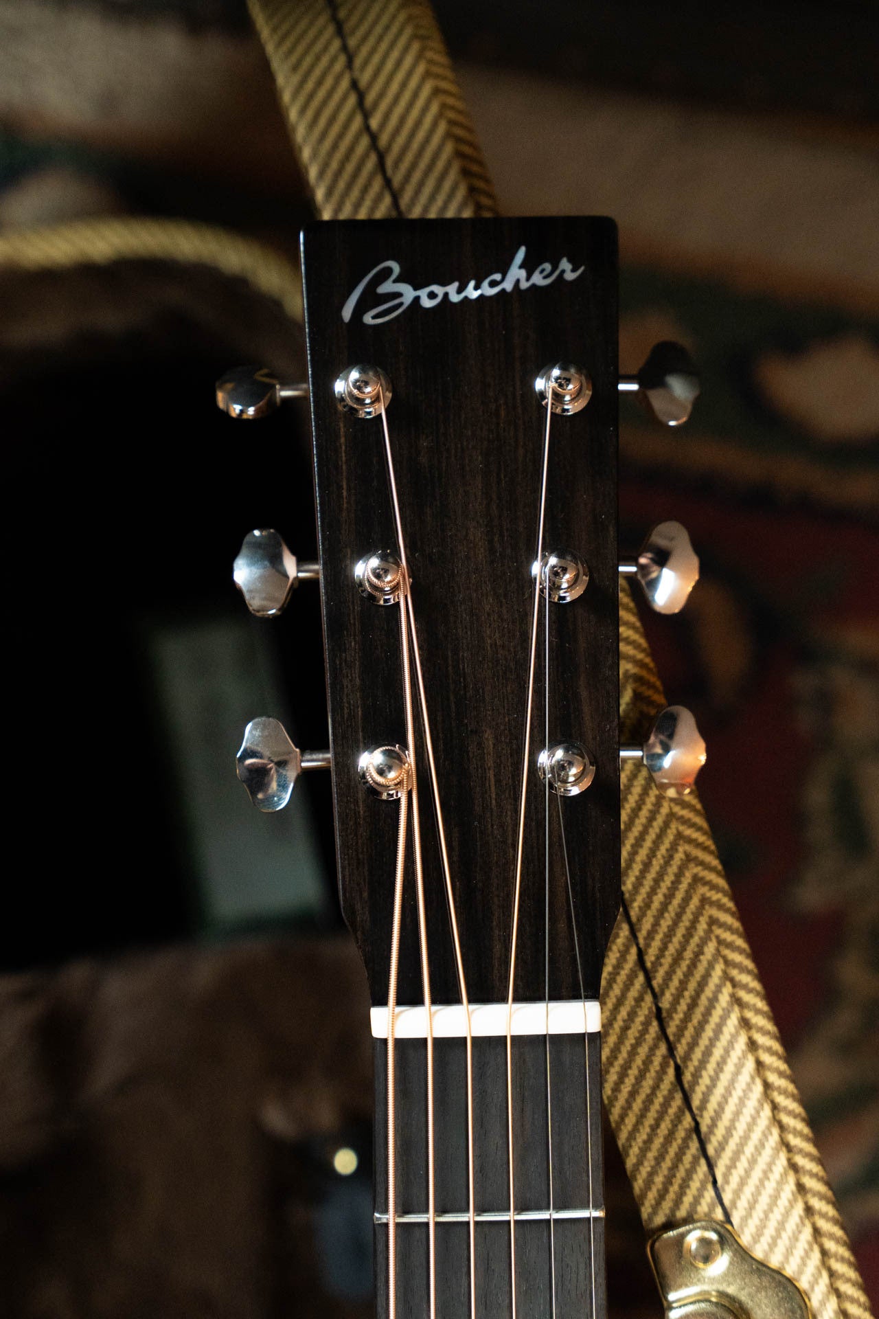 Close-up of a Boucher Guitars BG-42T-G Bluegrass Goose Series headstock with six tuners and a patterned strap. The All Torrefied Master Grade Adirondack Spruce/Mahogany build adds premium appeal. Case and fabric appear blurred in the background.
