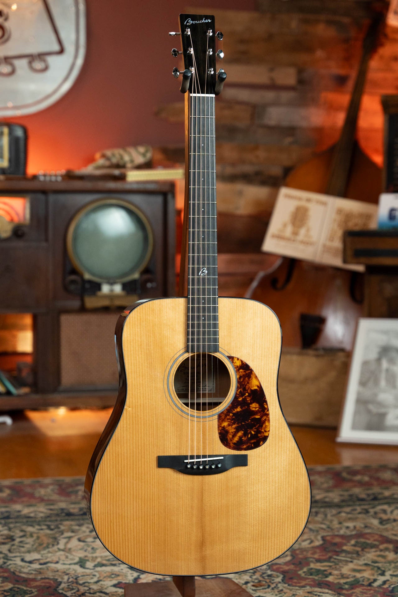 A Boucher Guitars BG-42T-G Bluegrass Goose Series Torrefied Adirondack/Mahogany Dreadnought Gold Pack Acoustic Guitar stands upright on a patterned rug, with vintage audio equipment and a double bass in the background.