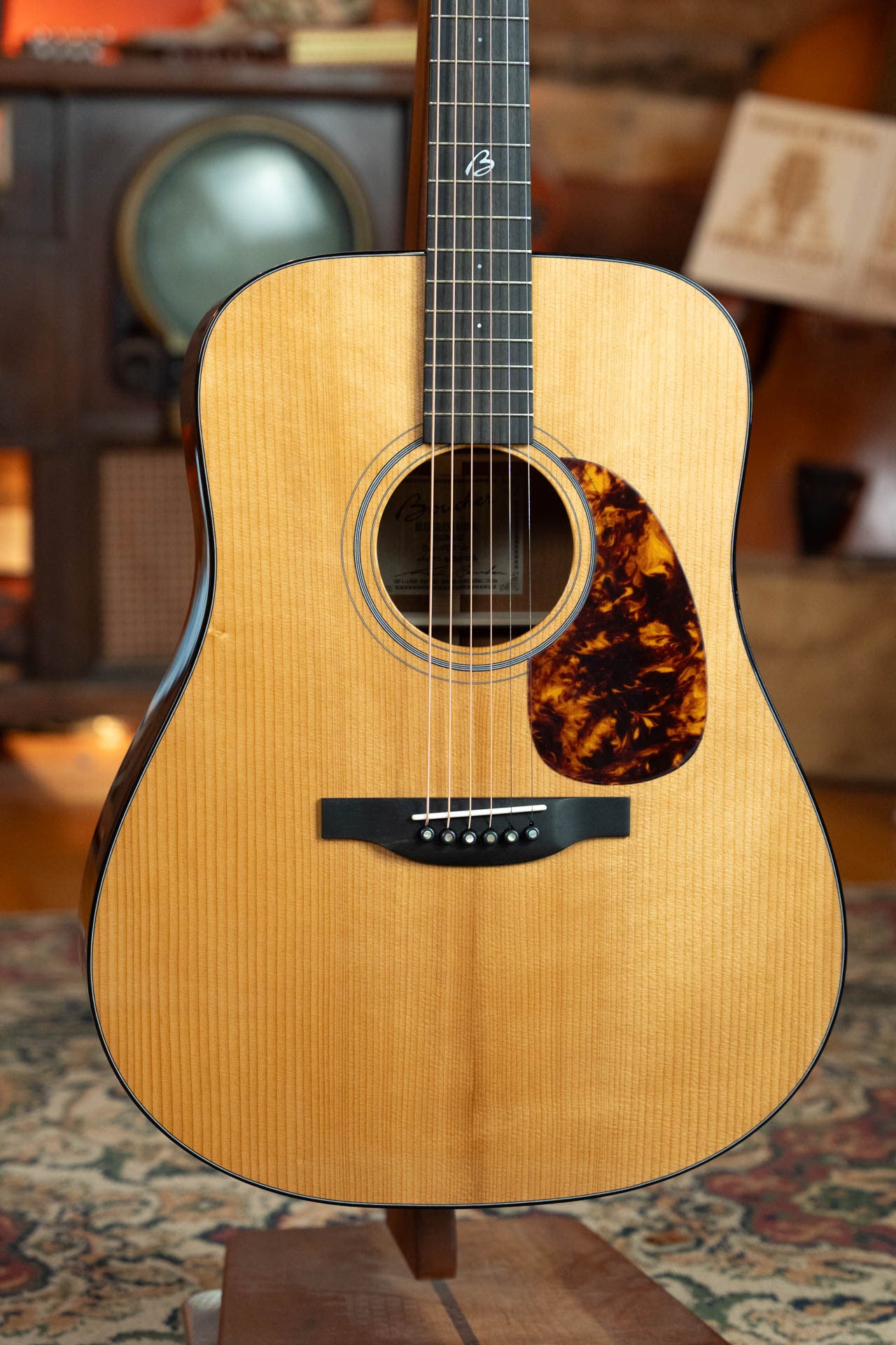 A Boucher Guitars BG-42T-G Bluegrass Goose Series *ALL* Torrefied Master Grade Adirondack/Mahogany Dreadnought Gold Pack Acoustic Guitar (#1035) with natural finish and tortoiseshell pickguard stands on a wooden stand in a cozy, vintage-decor room.