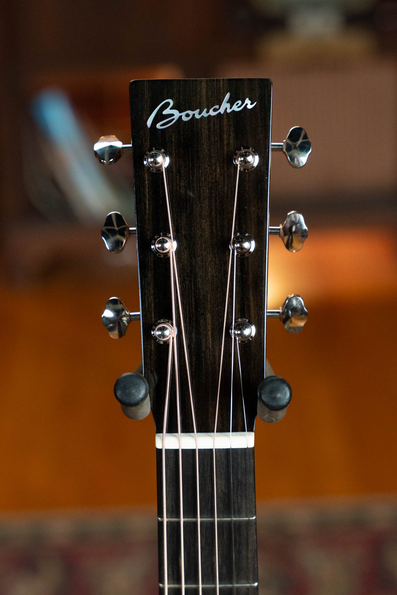 Close-up of the Boucher Guitars BG-42T-G Bluegrass Goose Series *ALL* Torrefied Master Grade Adirondack/Mahogany Dreadnought Gold Pack Acoustic Guitar headstock, with six tuning pegs and Boucher logo on a dark wood finish against a softly blurred warm background.