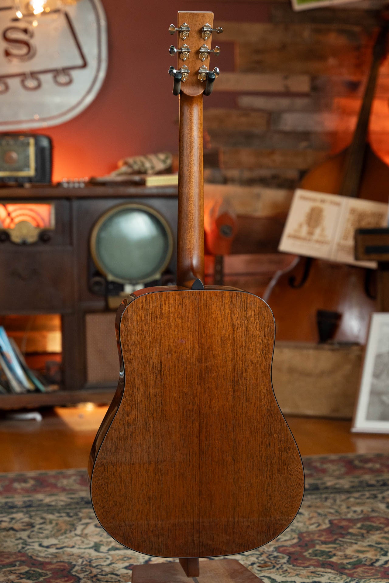 The Boucher BG-42T-G Bluegrass Goose Dreadnought by Boucher Guitars is shown from the back on a stand in a cozy, vintage-themed room with an old TV, double bass, and warm lighting.