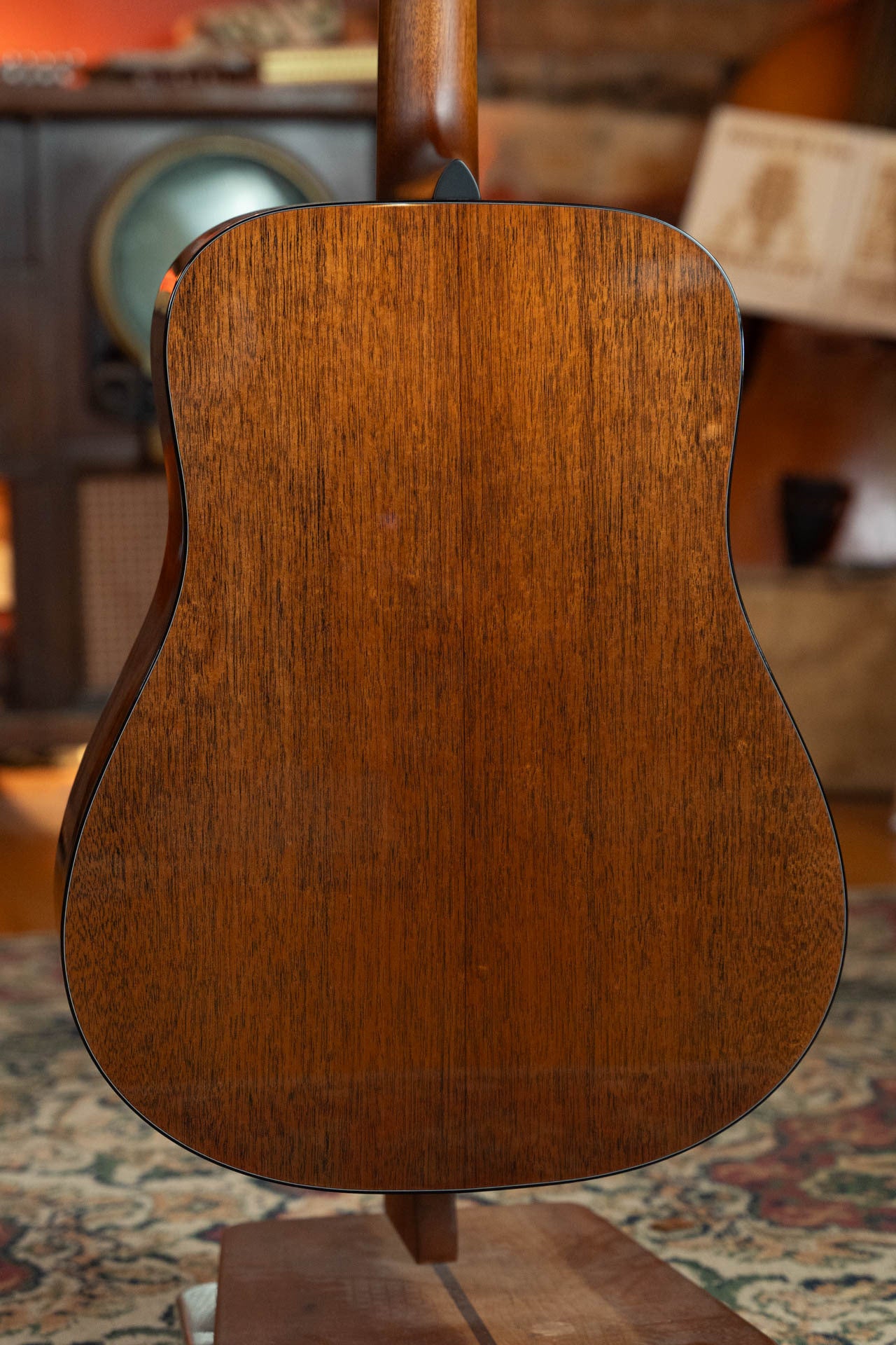 The image shows the back of a Boucher Guitars BG-42T-G Bluegrass Goose Series Torrefied Master Grade Adirondack/Mahogany Dreadnought Gold Pack Acoustic Guitar, upright indoors with blurred vintage furniture in the background.