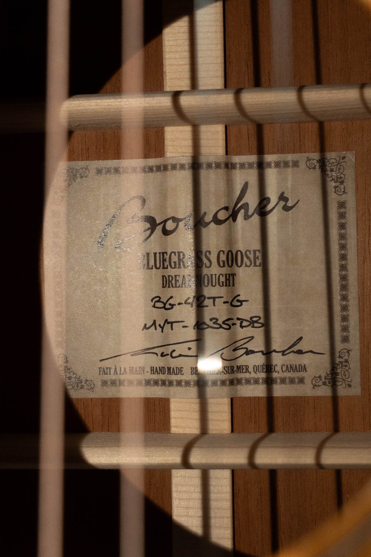 Close-up of a Boucher Guitars BG-42T-G Bluegrass Goose Series Dreadnought label (#1035) inside the soundhole, partially hidden by strings and shadows. Features all torrefied master grade Adirondack spruce and mahogany construction.