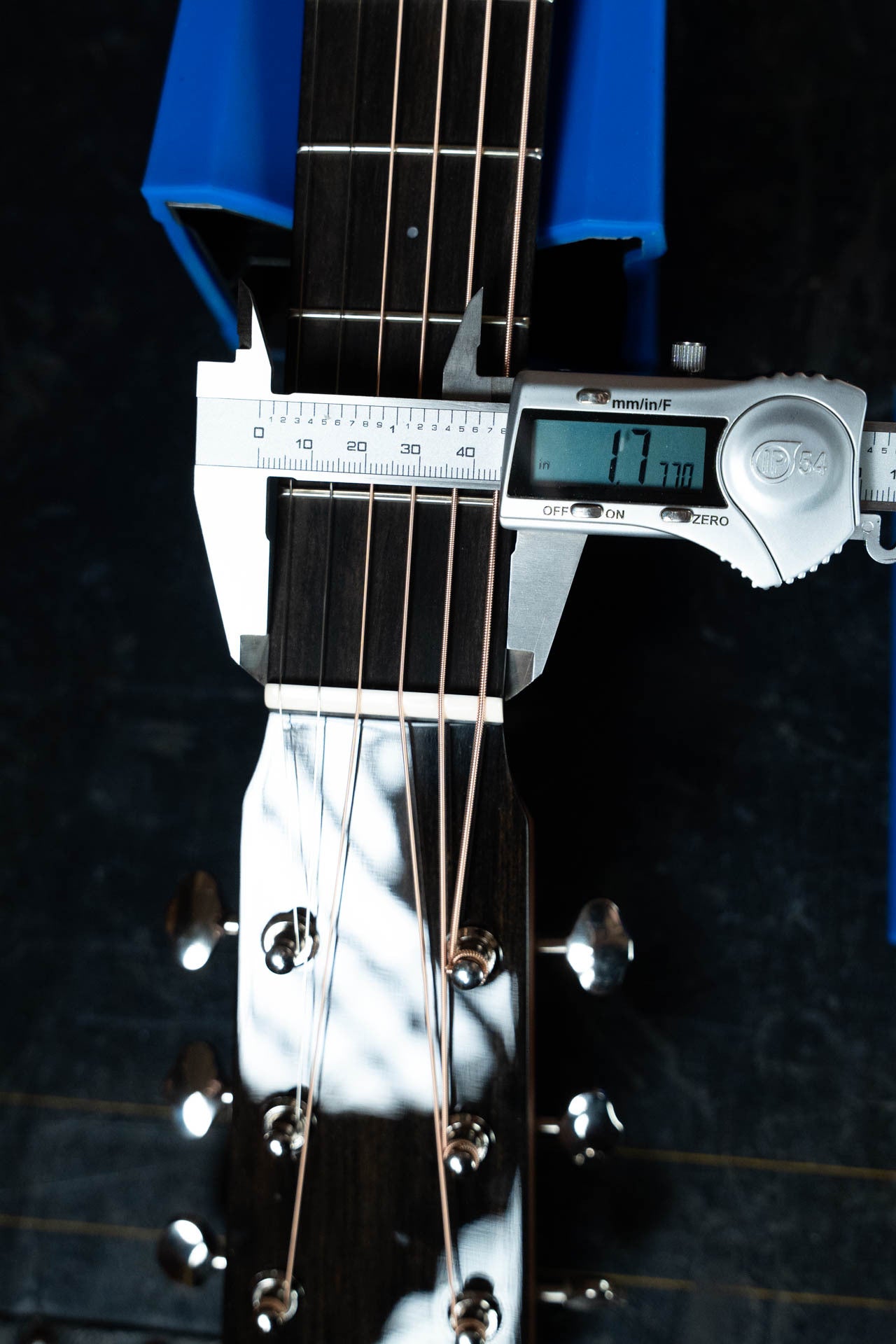 A digital caliper measures the nut width of a Boucher Guitars BG-42T-G Bluegrass Goose Series Dreadnought Acoustic Guitar #1035 at 47.70 mm, with the instrument resting in a blue holder and its strings and tuning pegs visible.