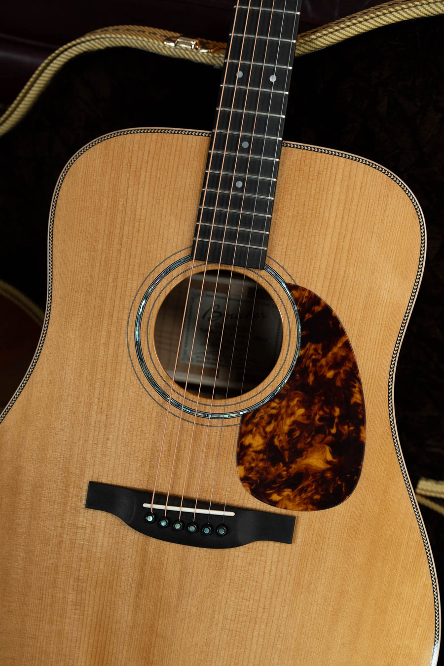 Close-up of a Boucher GR-SG-162T Grande Reserve dreadnought acoustic guitar (#1037) by Boucher Guitars, featuring the sound hole, pickguard, and bridge; the neck and part of a hardshell case are visible in the background.