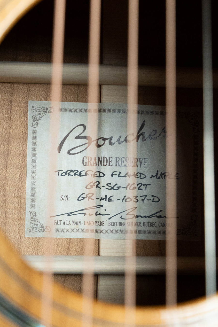 A close-up of a Boucher Guitars GR-SG-162T Grande Reserve Dreadnought #1037 label inside the soundhole, showing handwritten model and serial numbers, with guitar strings visible in the foreground.