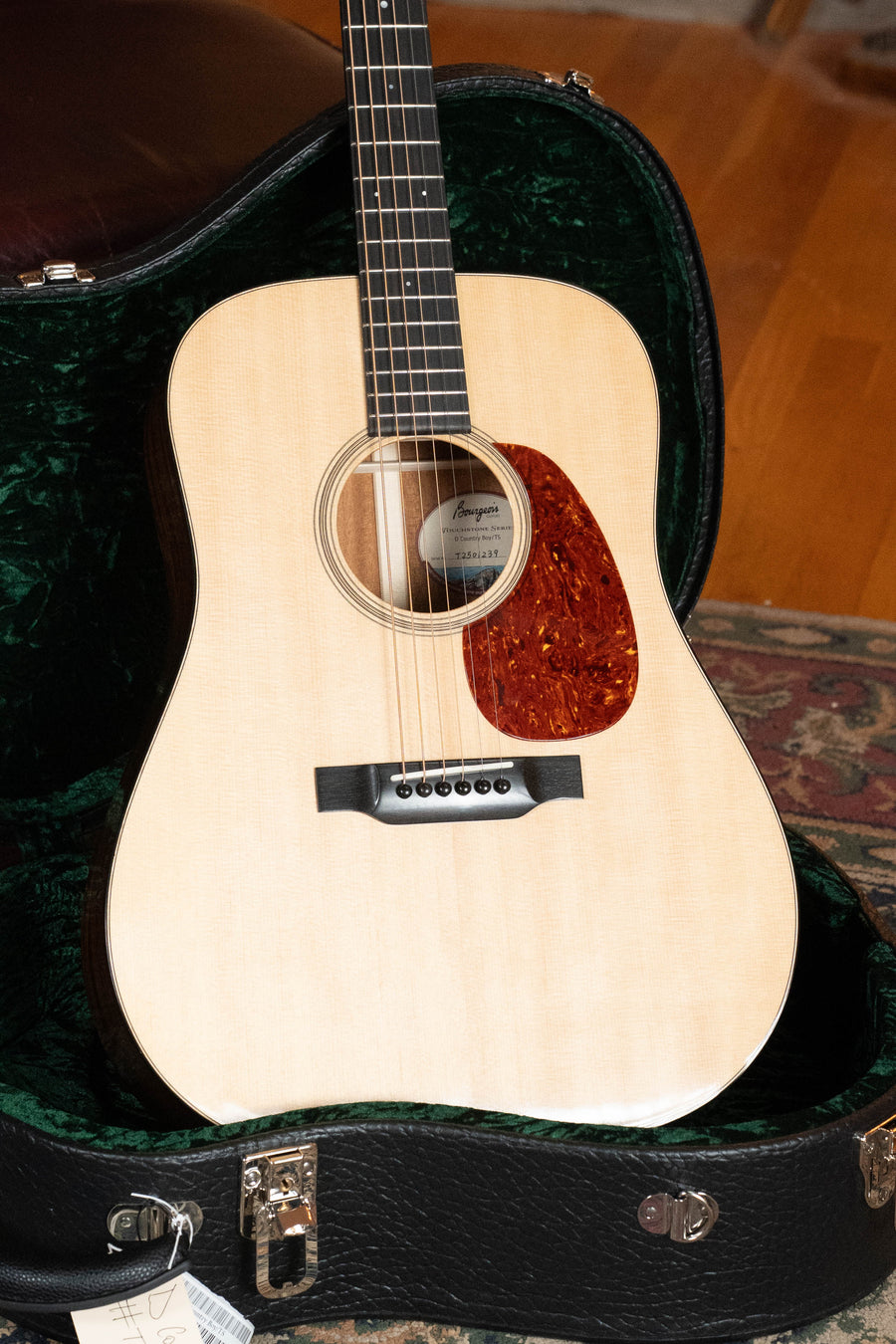 A Bourgeois Country Boy Touchstone Series Sitka/Mahogany Natural dreadnought acoustic guitar #1239 with a reddish-brown pickguard sits in an open, green-lined hard case on a wooden floor, featuring six strings and a tag on the case latch.