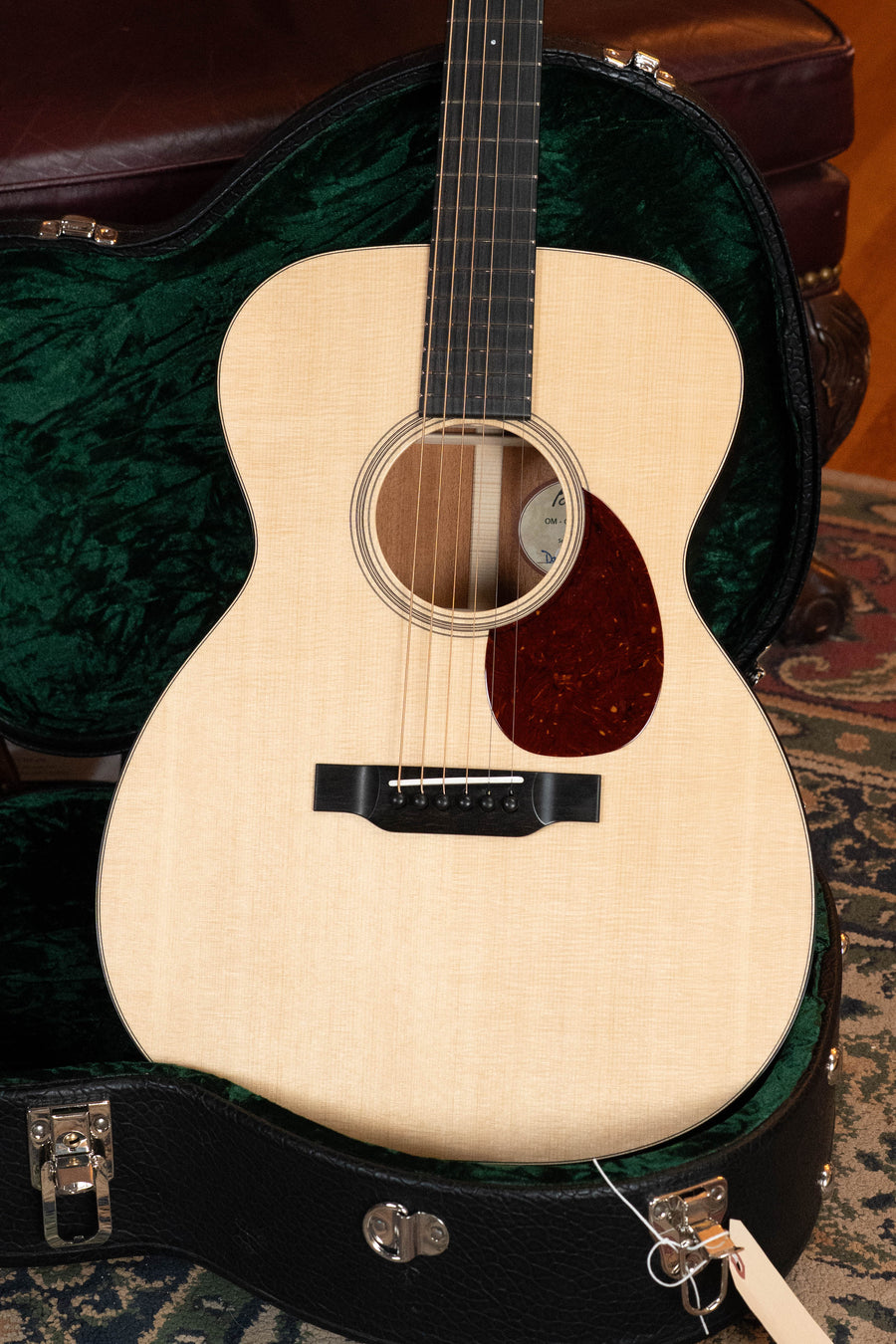 A Bourgeois Professional Series Country Boy Satin Sitka/Mahogany Satin OM Acoustic Guitar #1219 rests in an open black hard case with green plush lining, placed on a patterned rug beside a leather chair.
