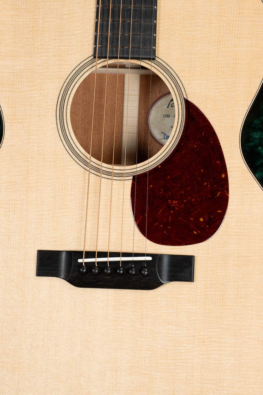 Close-up of the Bourgeois Professional Series Country Boy Satin Sitka/Mahogany OM Acoustic Guitar #1219, showing its six strings, wooden bridge, round sound hole with label, and dark red pickguard.