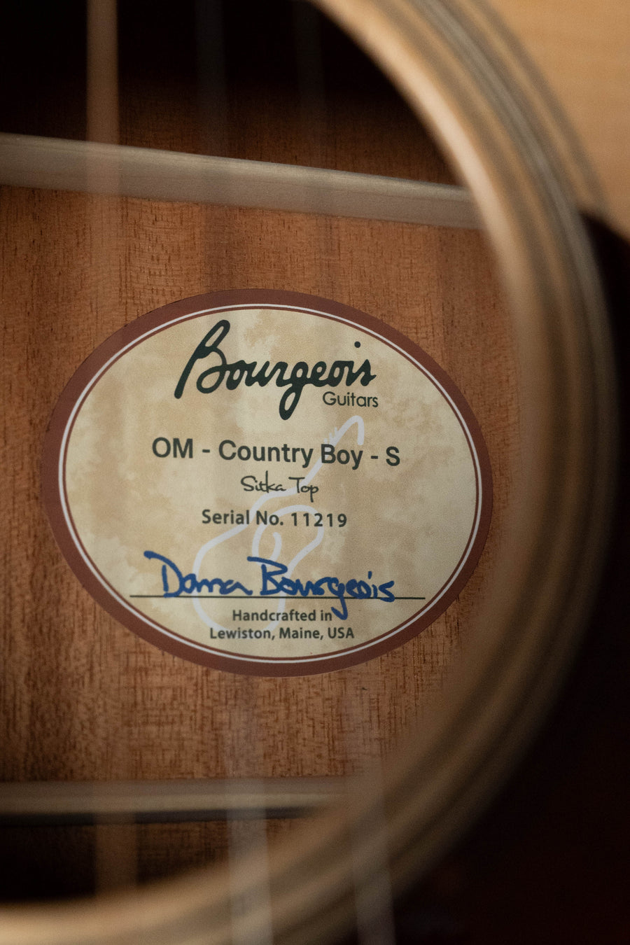 A close-up through the strings shows the inside label of a Bourgeois Professional Series Country Boy Satin Sitka/Mahogany OM Acoustic Guitar #1219, handcrafted in Lewiston, Maine, USA.
