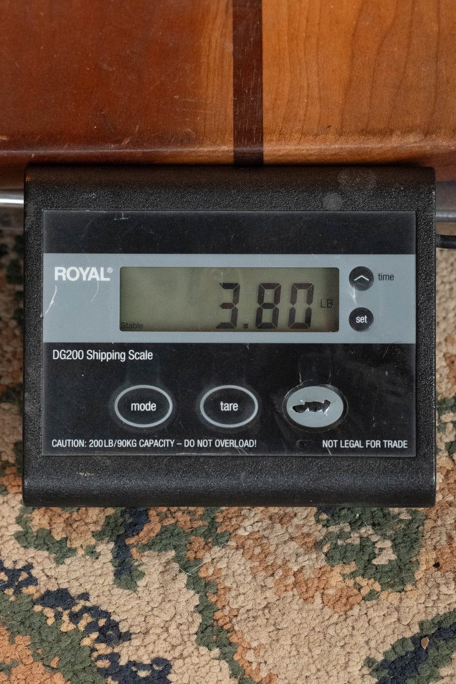 A digital shipping scale shows 3.80 pounds—the weight of your Bourgeois Professional Series Country Boy Satin Sitka/Mahogany OM Acoustic Guitar #1219—on a patterned carpet near wood. The scale has mode, tare, and set buttons.