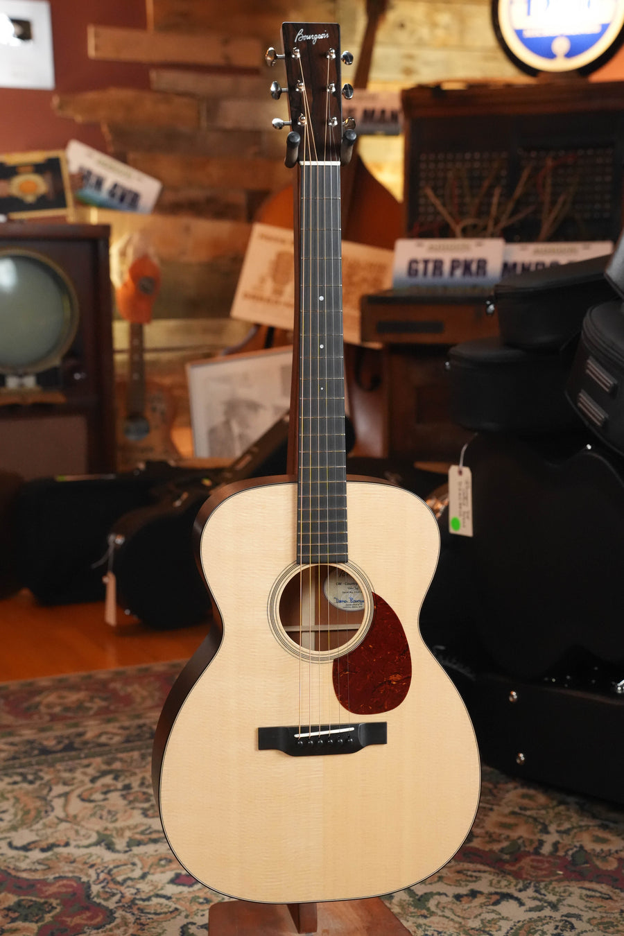 A Bourgeois Professional Series Country Boy Satin Sitka/Mahogany OM Acoustic Guitar (#1414) stands upright on a patterned rug in a cozy room filled with vintage music equipment and décor.