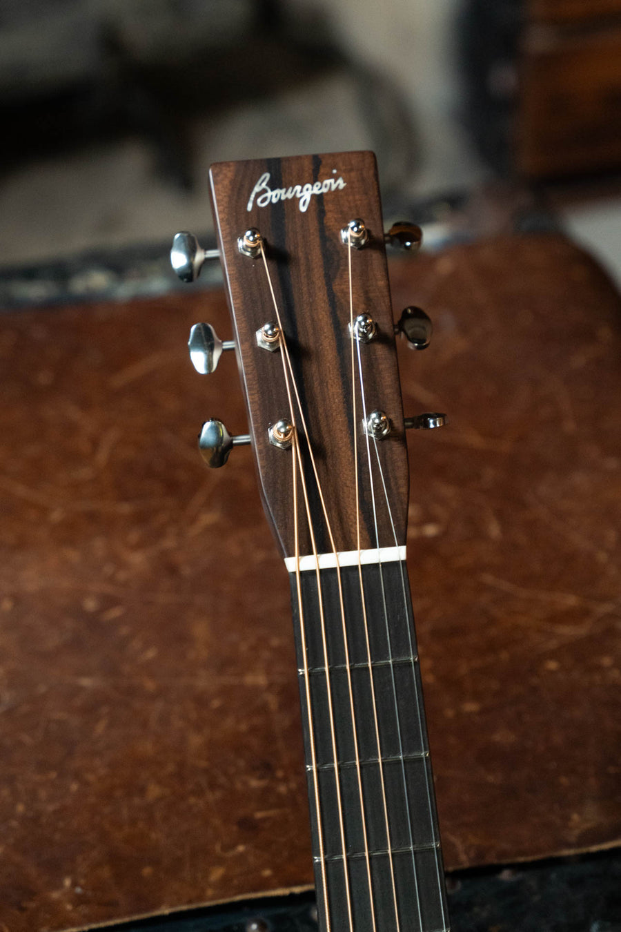 Close-up of a Bourgeois Touchstone Country Boy Sitka/Mahogany Natural Dreadnought Acoustic Guitar #8237 headstock, featuring the Bourgeois logo on dark wood, silver tuning pegs, and six strings against a blurred brown background.