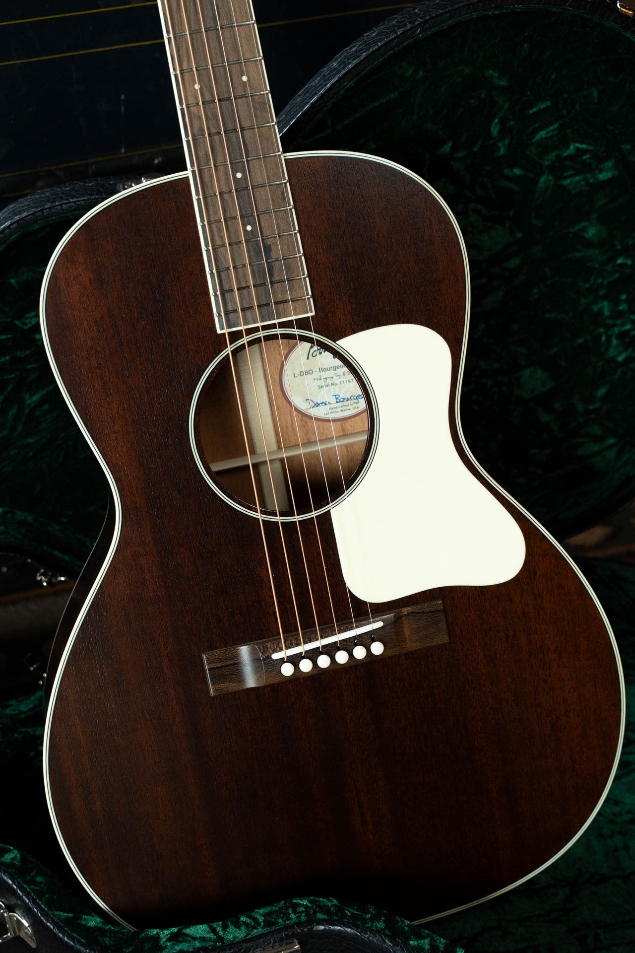 A close-up of a Bourgeois Blues PSBB-LDBO-S Satin Professional Series All Mahogany OO Acoustic Guitar #1197, featuring a dark wood finish, white pickguard, and six strings, resting in an open green-lined hard case.