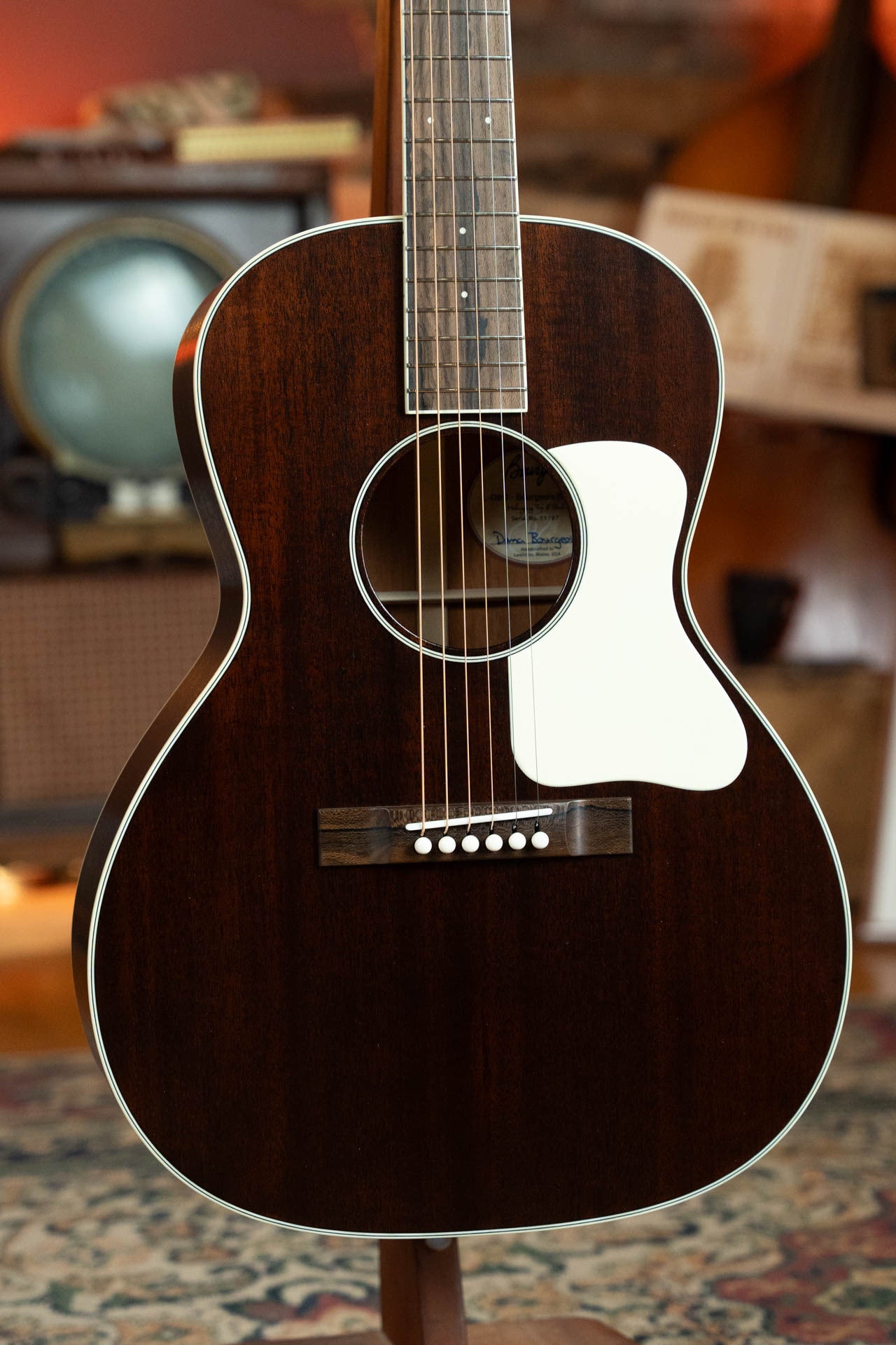 A close-up of a Bourgeois Blues PSBB-LDBO-S Satin Professional Series All Mahogany OO Acoustic Guitar #1197 with a dark wood finish, white pickguard, and six strings stands on a patterned rug, vintage TV and books blurred in the background.