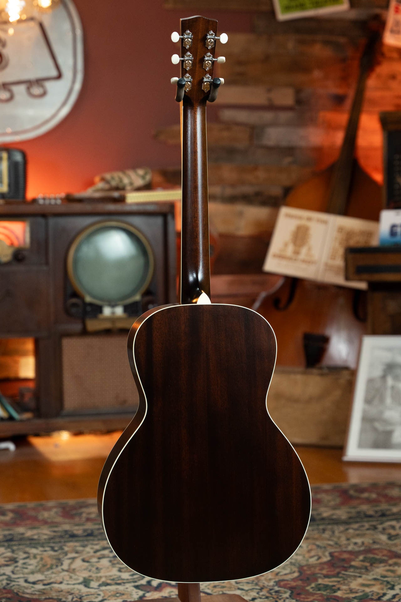 A Bourgeois Blues PSBB-LDBO-S Satin Professional Series All Mahogany OO Acoustic Guitar #1197 stands upright, shown from the back in a warmly lit, vintage-inspired room with a double bass, old radio, and framed pictures visible.