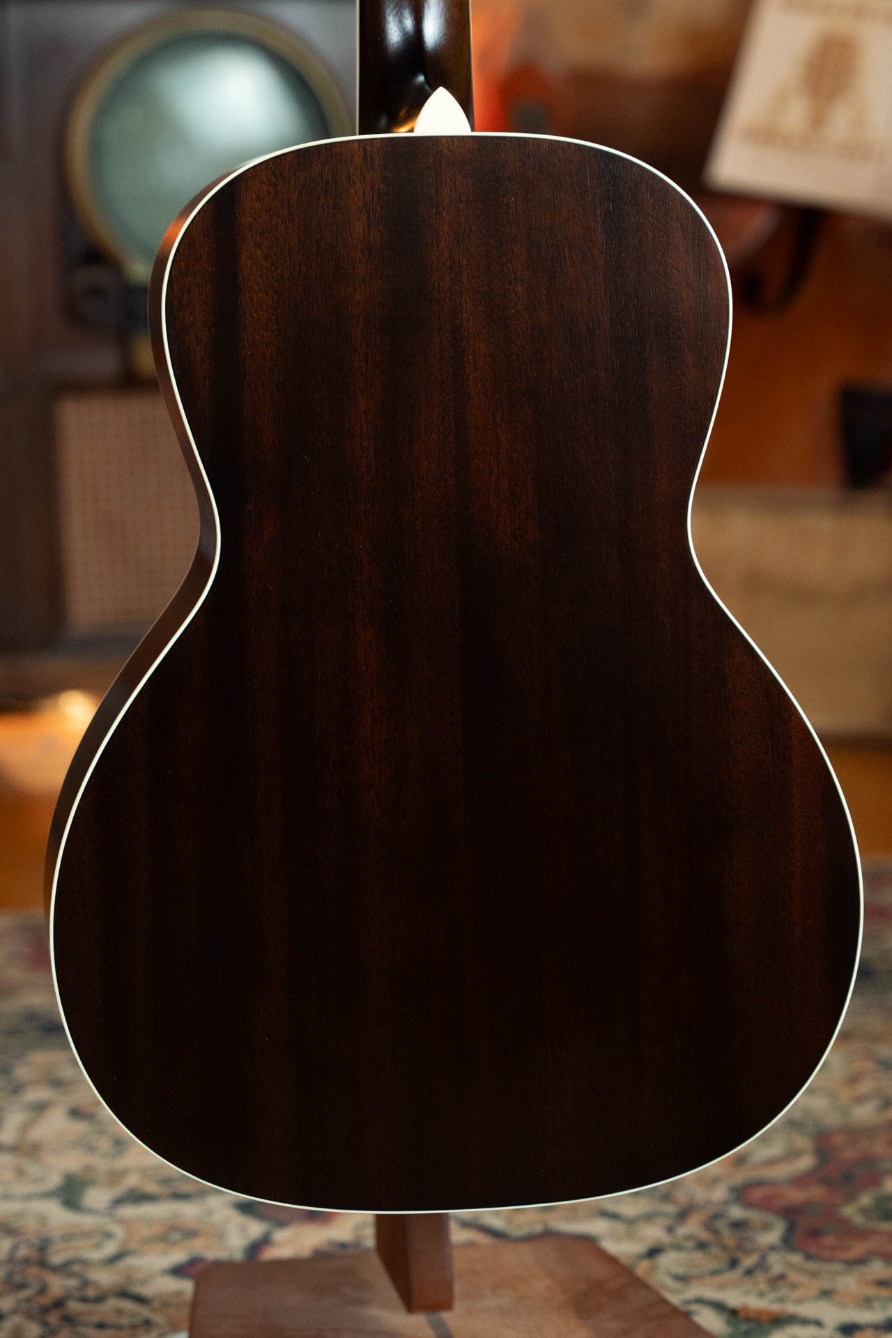 The image shows the back of a Bourgeois Blues PSBB-LDBO-S Satin Professional Series All Mahogany OO Acoustic Guitar #1197, displayed upright on a stand with dark wood grain and white binding. A softly blurred background features vintage decor and a patterned rug.