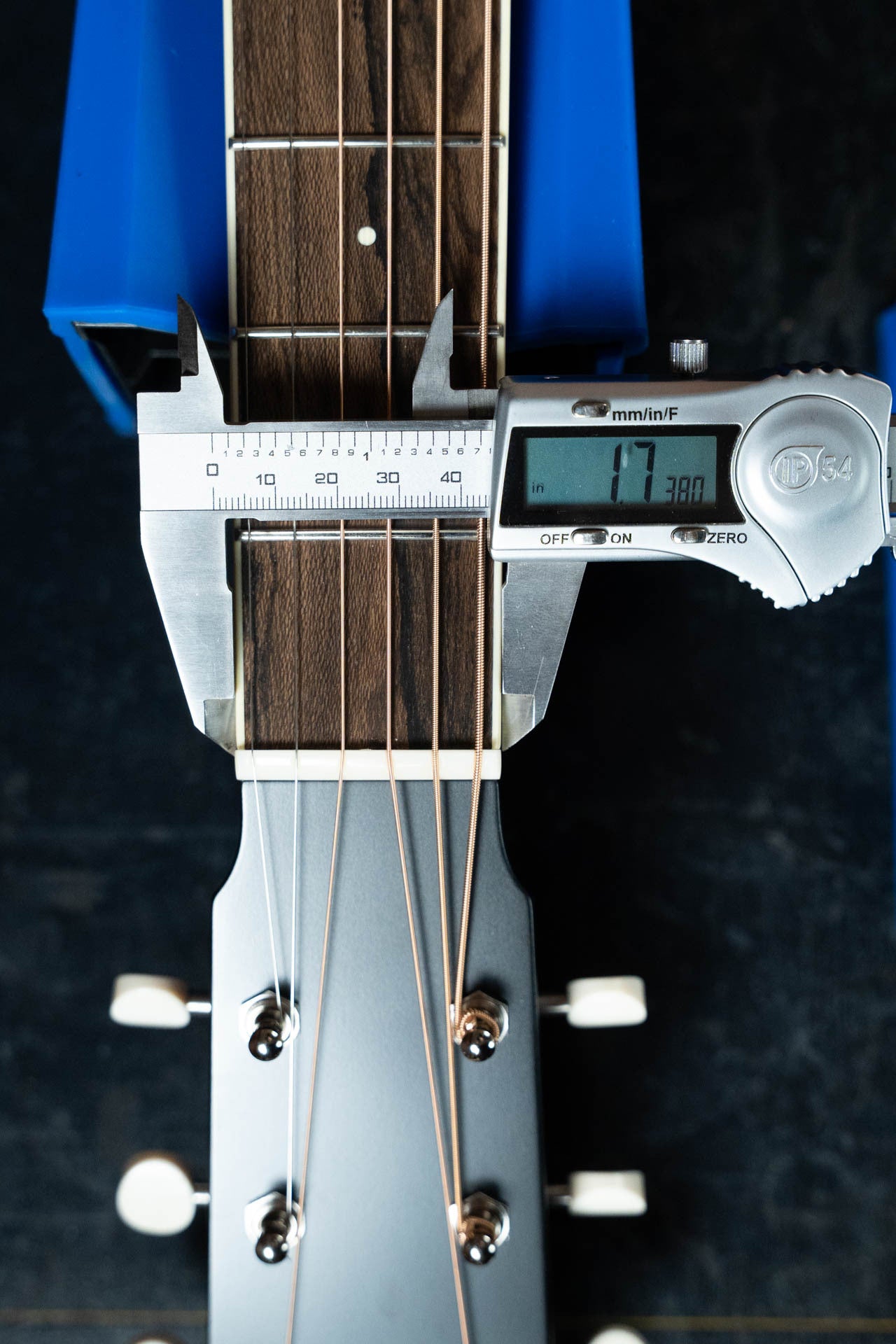A digital caliper measures the width of a Bourgeois Blues PSBB-LDBO-S Satin Professional Series All Mahogany OO Acoustic Guitar #1197 neck near the headstock, displaying 47.38 mm on its screen, with strings and tuning pegs visible against a dark background.