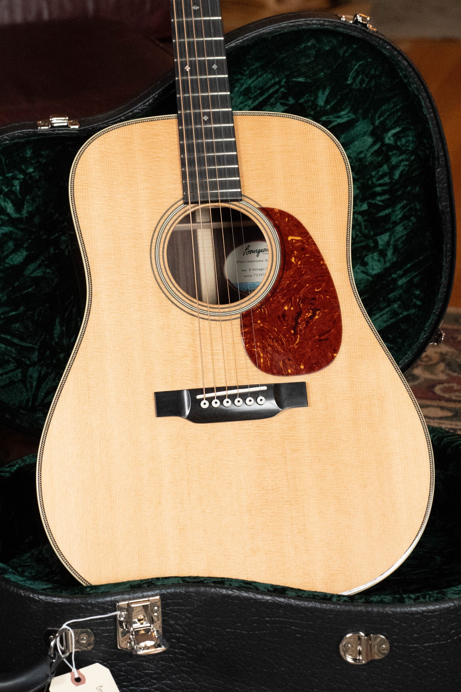 A Bourgeois D Vintage/TS Touchstone Series Dreadnought Acoustic Guitar #7014 with a tortoiseshell pickguard rests in an open black hard case lined with green velvet, showing its latch and tag at the bottom. Crafted from Old-Growth Alaskan Sitka Spruce.