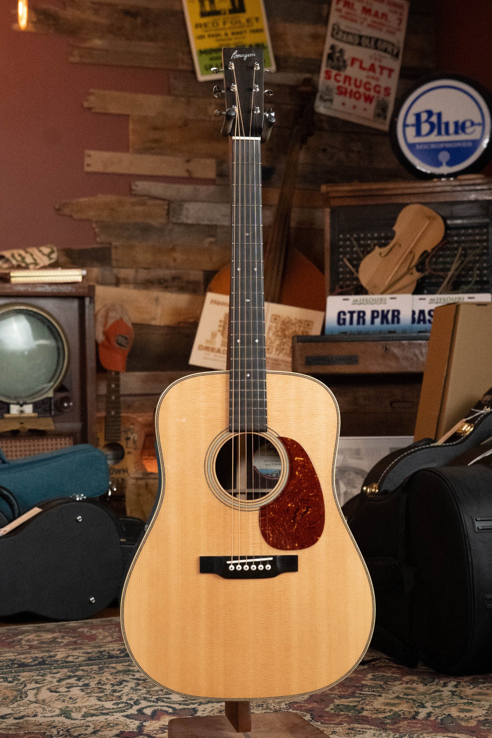 A Bourgeois D Vintage/TS Touchstone Series Dreadnought Acoustic Guitar #7014 by Bourgeois stands upright on a stand in a cozy room filled with vintage posters, a TV, and musical instruments.