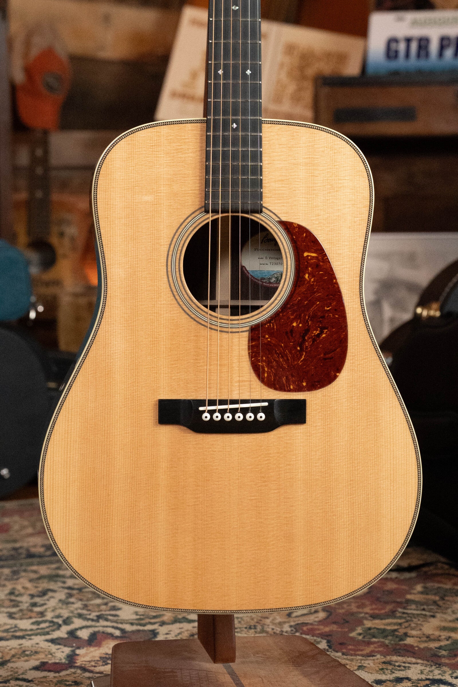 A close-up of the Bourgeois D Vintage/TS Touchstone Series Dreadnought Acoustic Guitar #7014, featuring an Old-Growth Alaskan Sitka Spruce top and dark pickguard, displayed upright on a stand indoors with musical gear in the background.