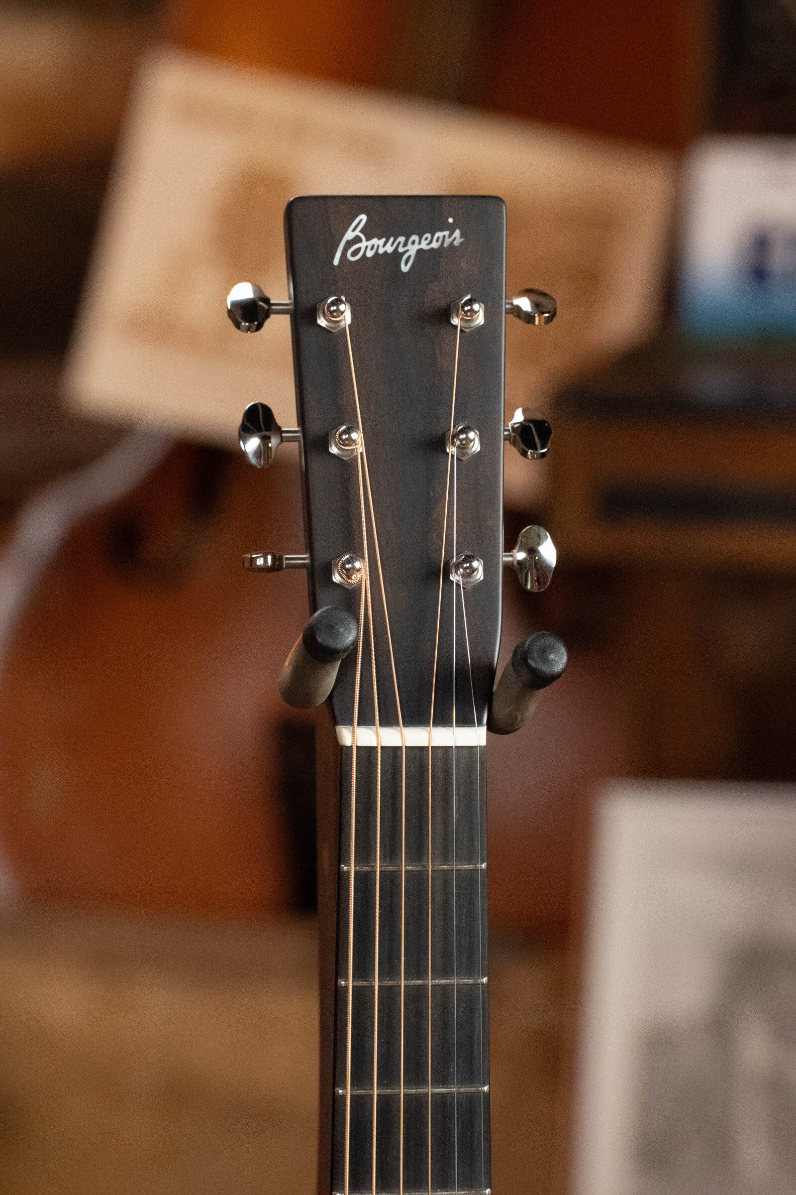 Close-up of the headstock on the Bourgeois D Vintage/TS Touchstone Series Dreadnought Acoustic Guitar #7014, featuring tuning pegs and the Bourgeois logo, with a blurred background of wood and other objects.