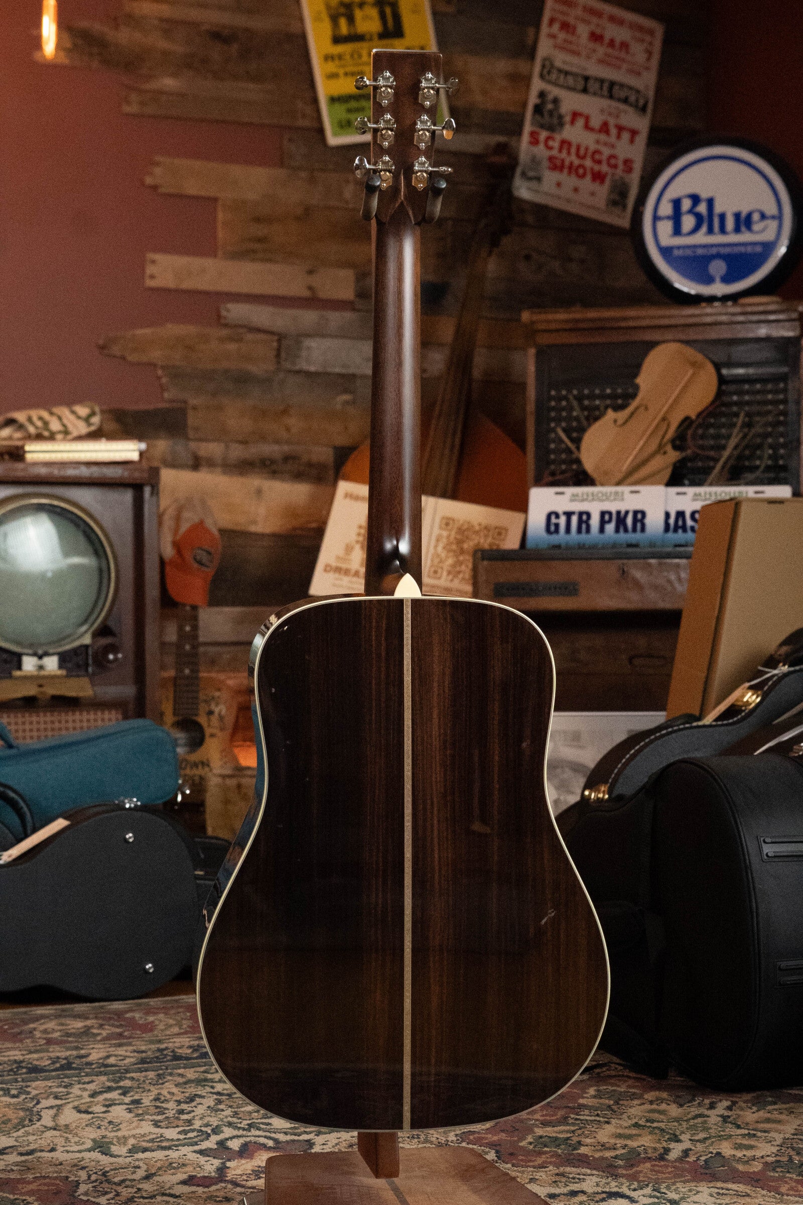 A Bourgeois D Vintage/TS Touchstone Series Dreadnought Acoustic Guitar #7014 stands upright, back view showing its dark, glossy finish in a cozy room filled with vintage music posters, amplifiers, and a patterned rug.